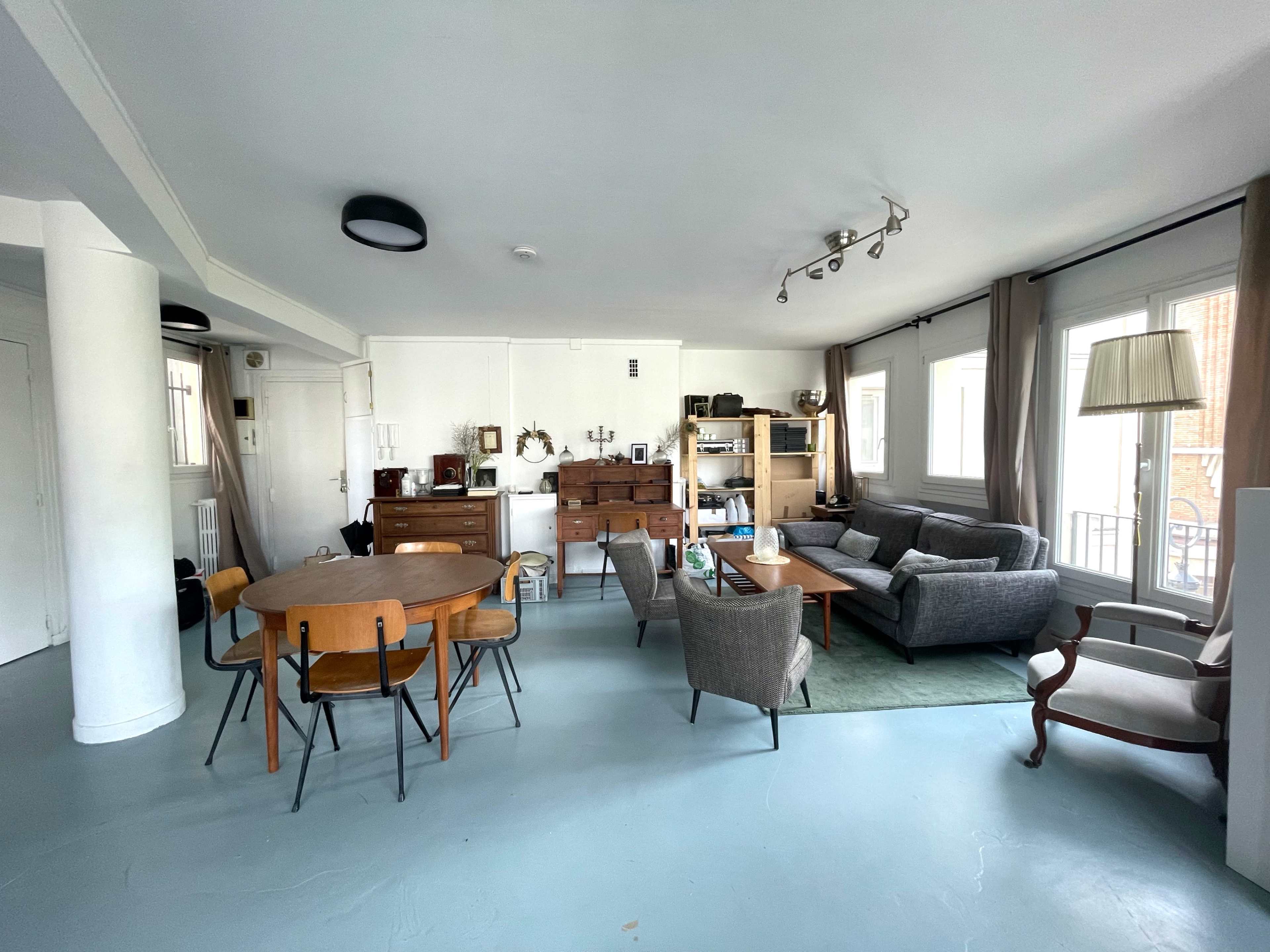 The image shows a spacious living room furnished with a gray sofa, wooden dining table, and various shelves displaying decorative items.