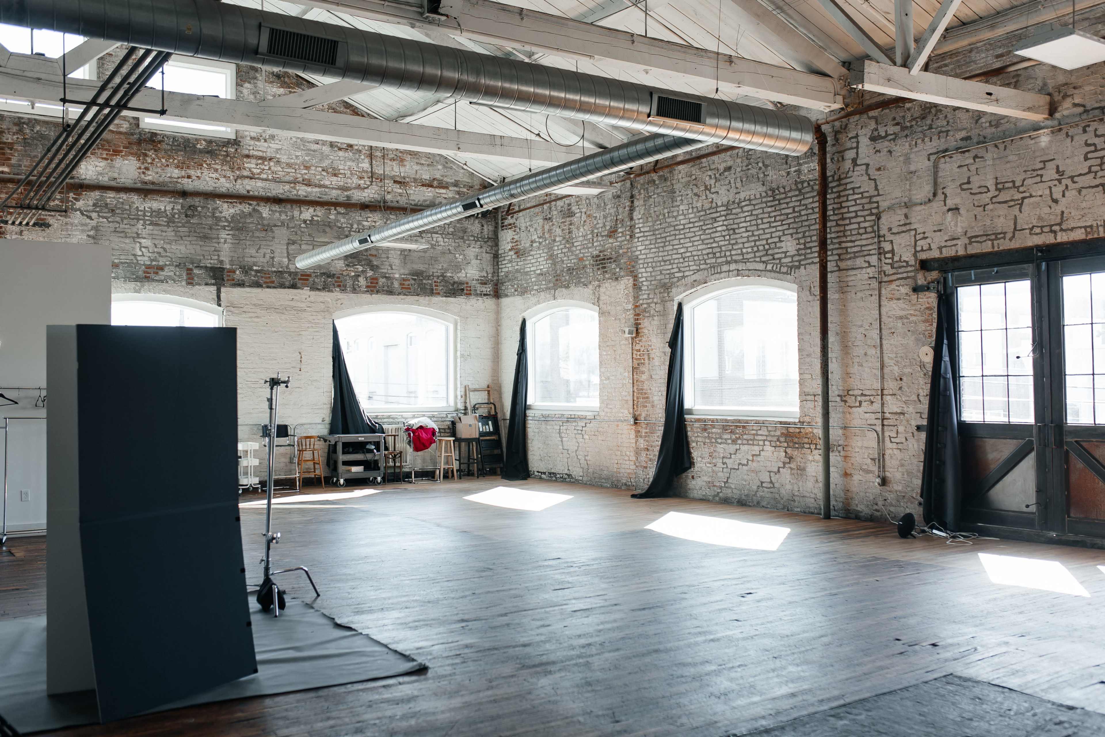 An empty studio space with exposed brick walls, large windows, and wooden floors, featuring a backdrop and lighting equipment set up in one corner.