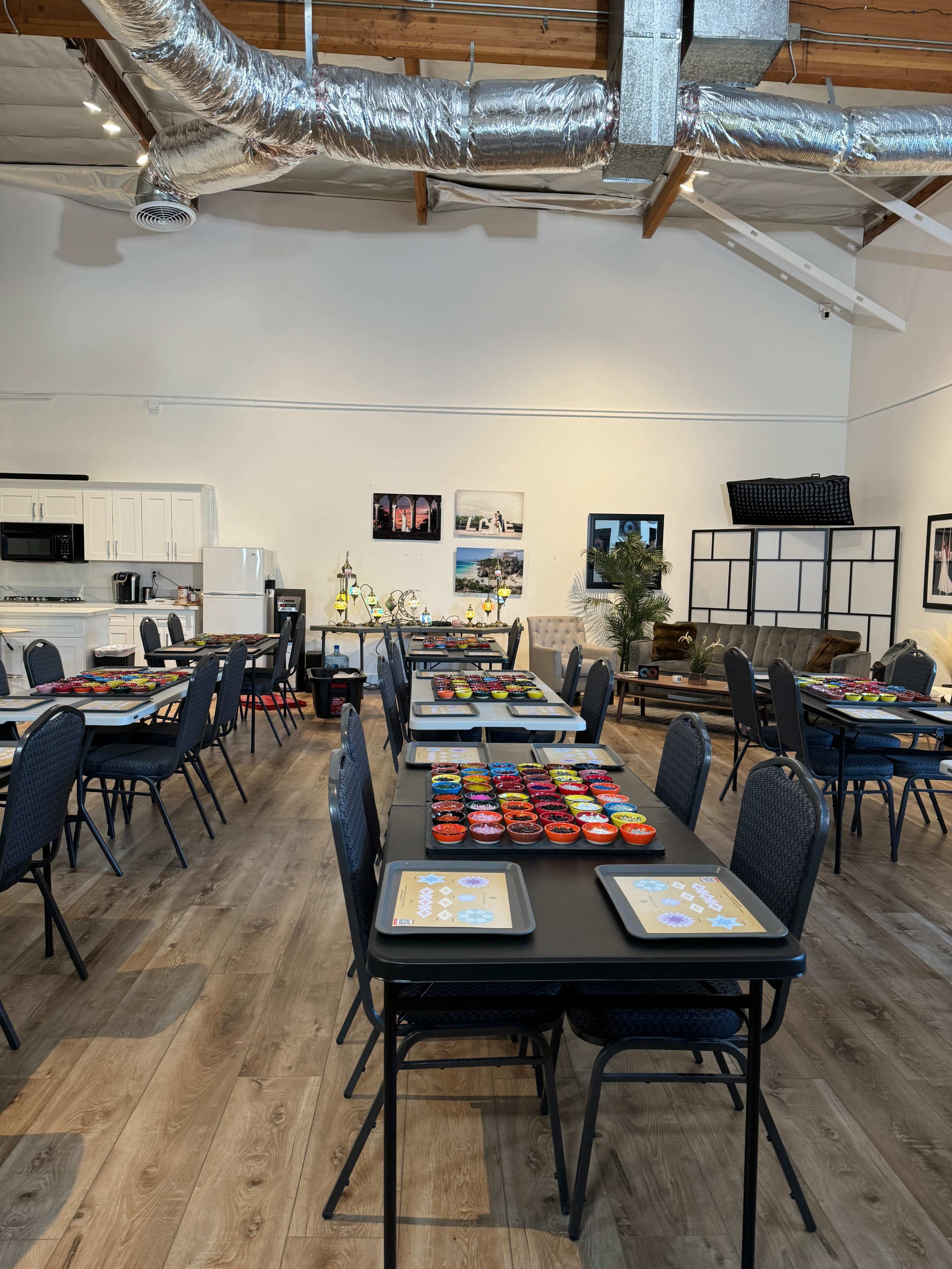 An interior space features multiple tables set with colorful dishes, a kitchenette in the background, and framed photographs on the walls.