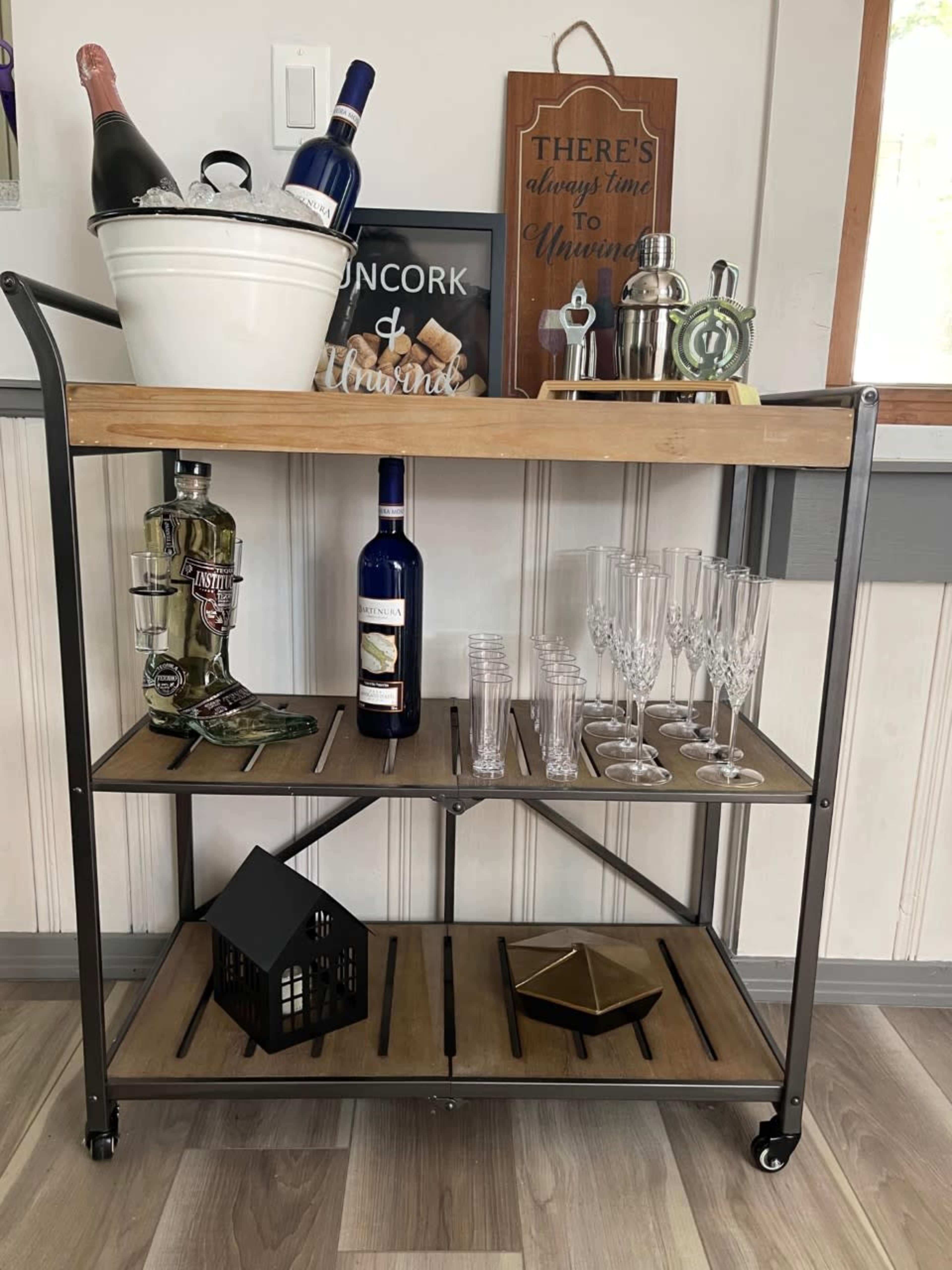 A wooden bar cart displays various bottles of alcohol, glassware, and a decorative item against a wall with a framed sign.