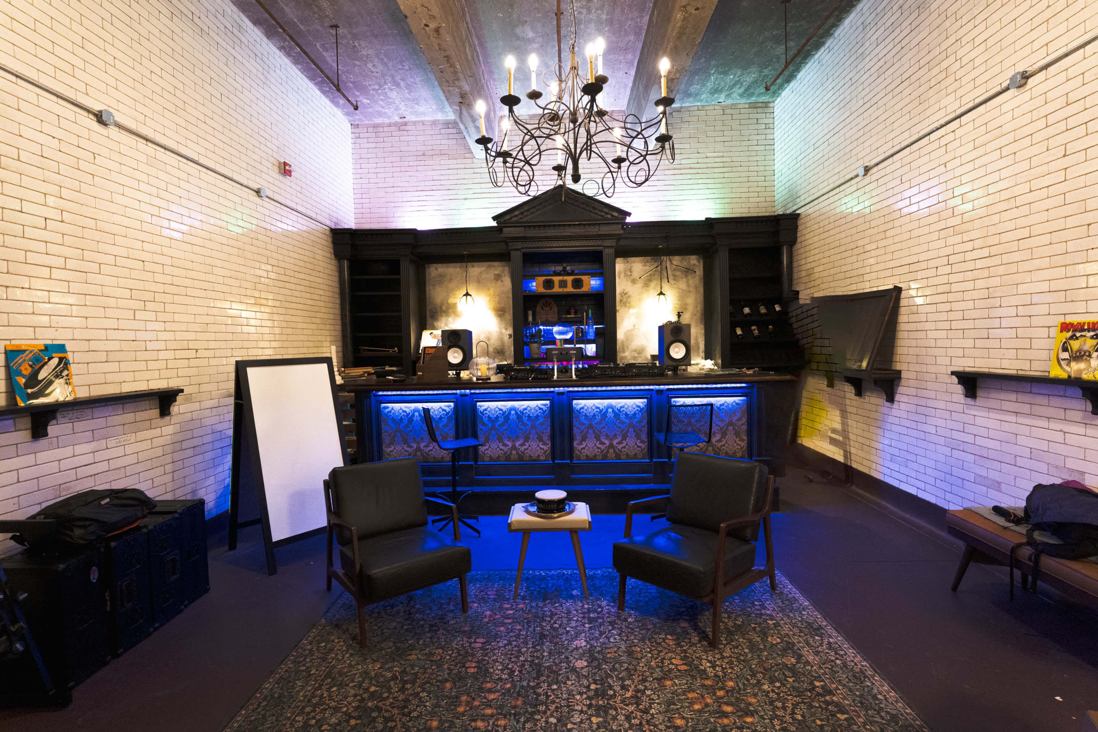 The image shows a modern bar area with a chandelier, two chairs, a small table, and a wall covered in white tiles, illuminated by colored lights.