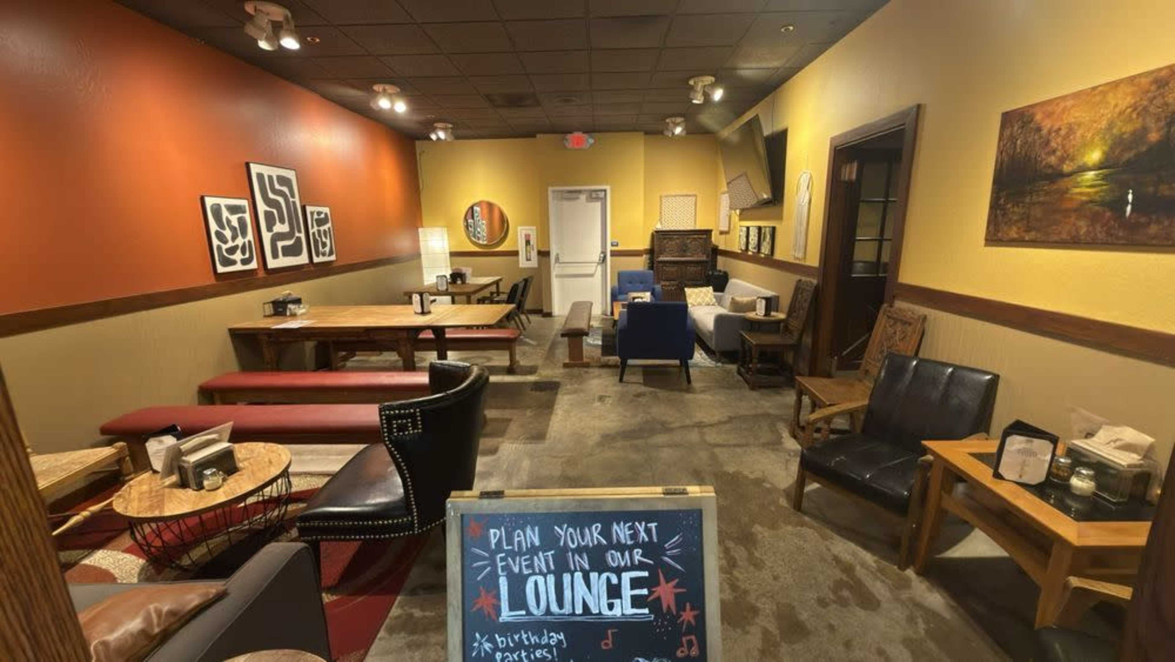 The image shows a cozy lounge with various seating options, including wooden tables, couches, and chairs, along with a chalkboard promoting event bookings.
