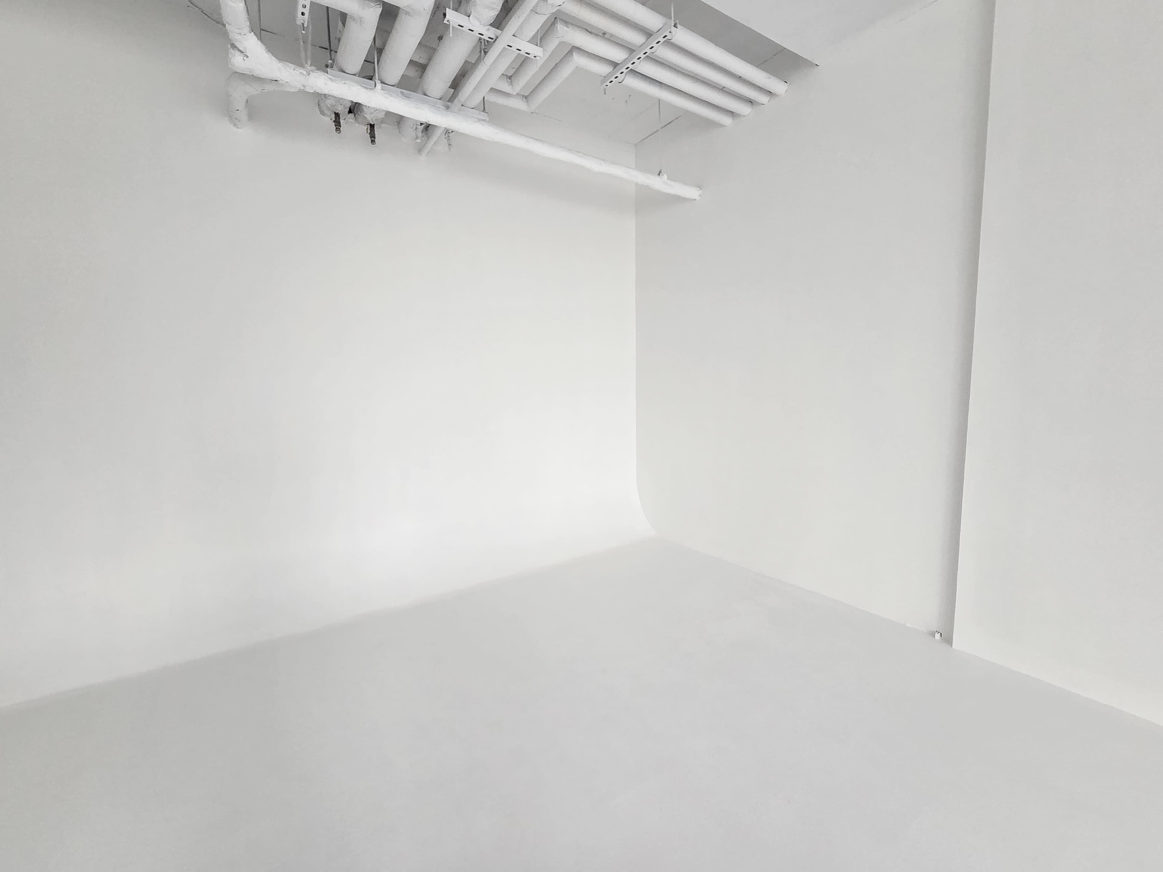 The image shows a spacious, empty room with white walls and a white floor, featuring exposed pipes on the ceiling.