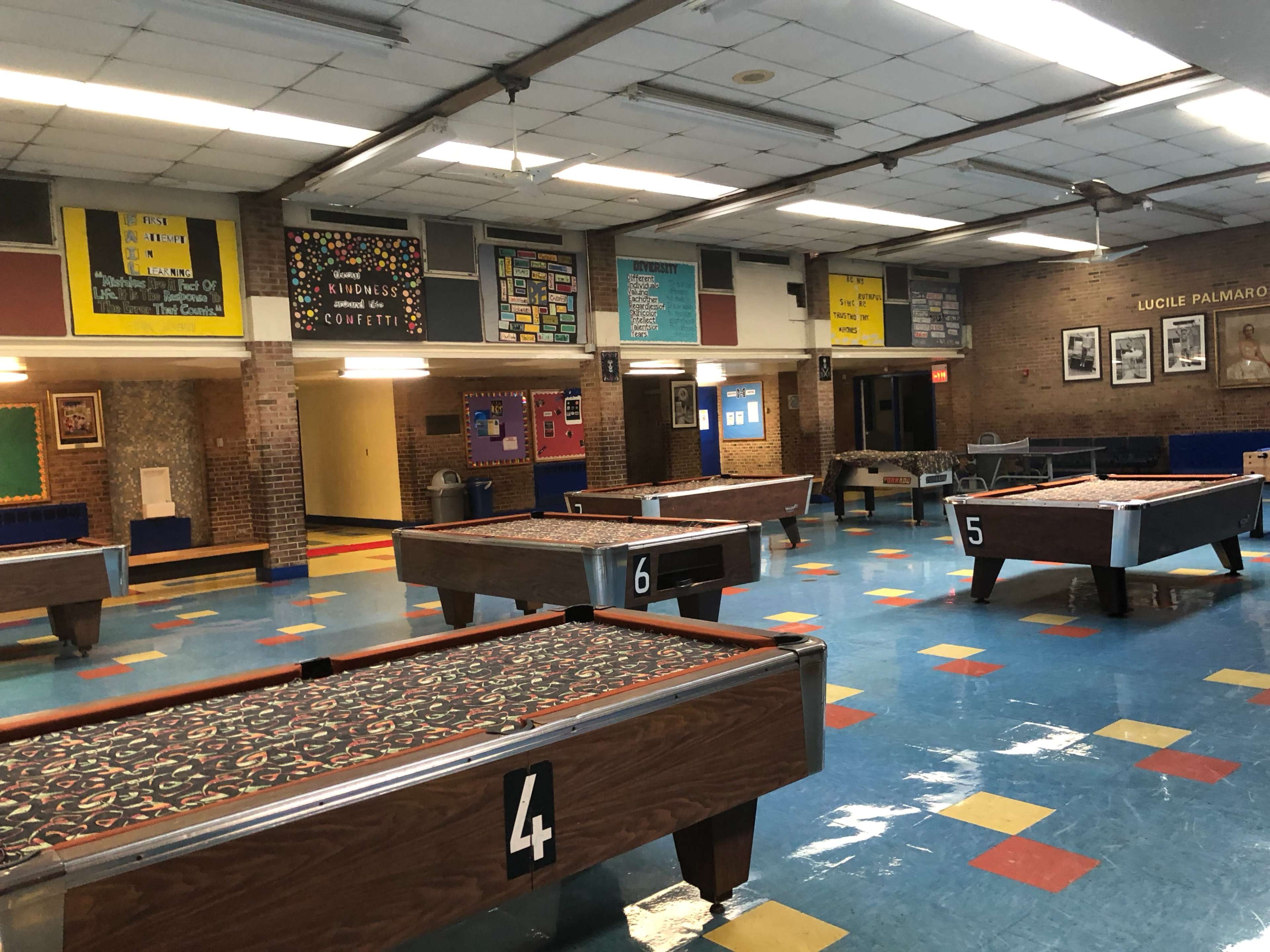 An empty gymnasium with several pool tables labeled with numbers, colorful floor tiles, and bulletin boards on the walls.