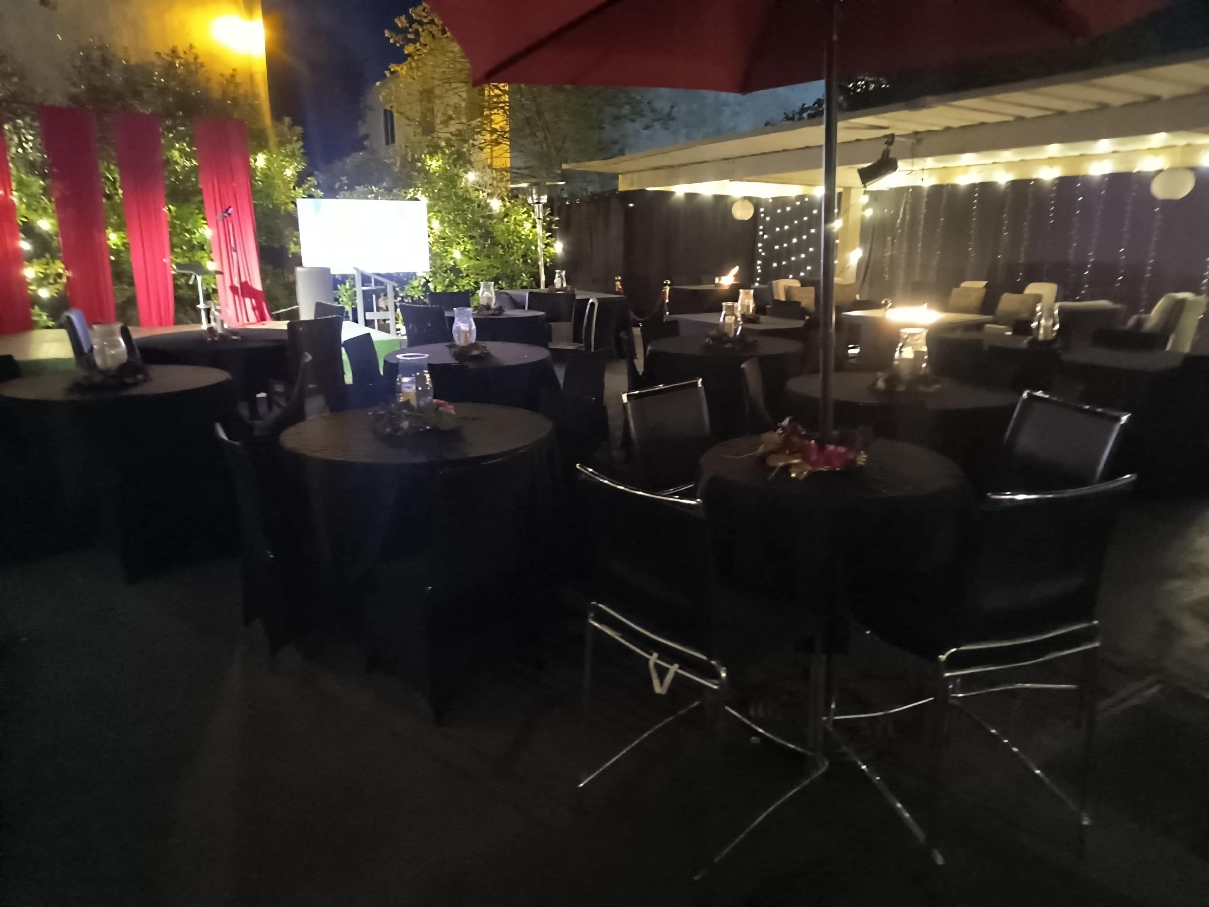 The image shows a dimly lit outdoor event space with several round tables covered in black tablecloths, surrounded by decorative lights and an umbrella.