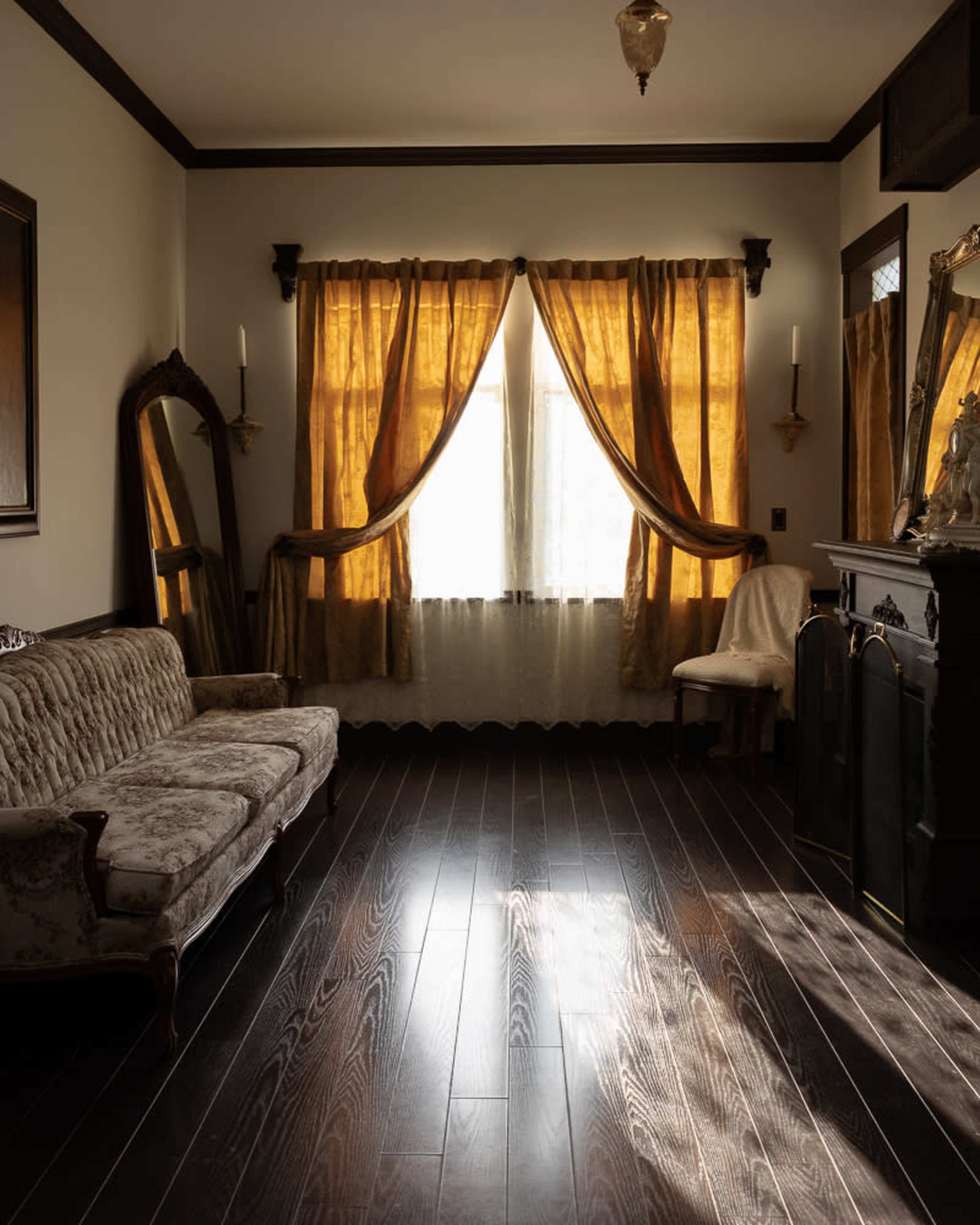 The image shows a room with dark wooden flooring, a vintage couch, and large windows draped with golden curtains.