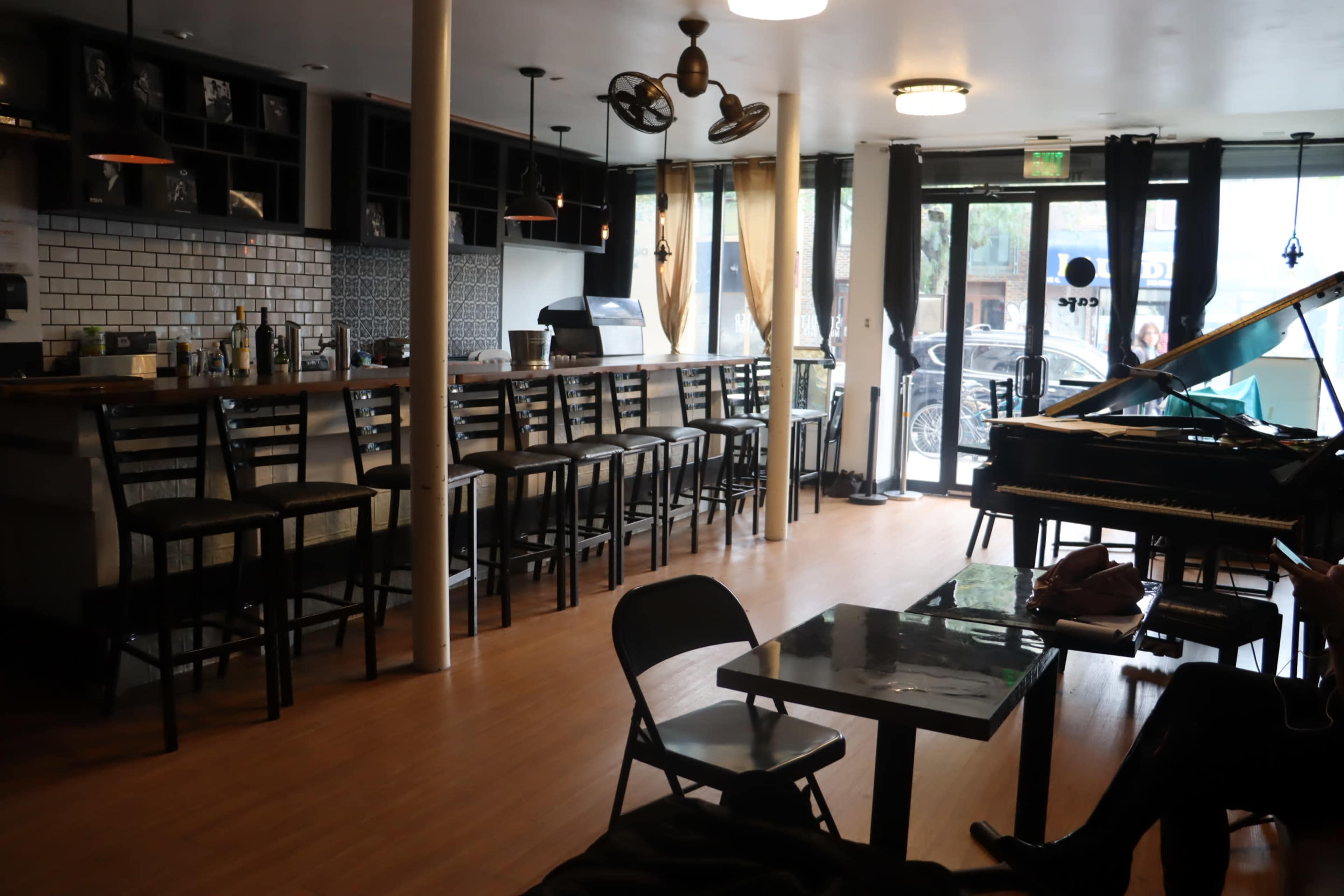 A spacious café interior with a long counter, bar stools, a piano, and large windows allowing natural light to enter.