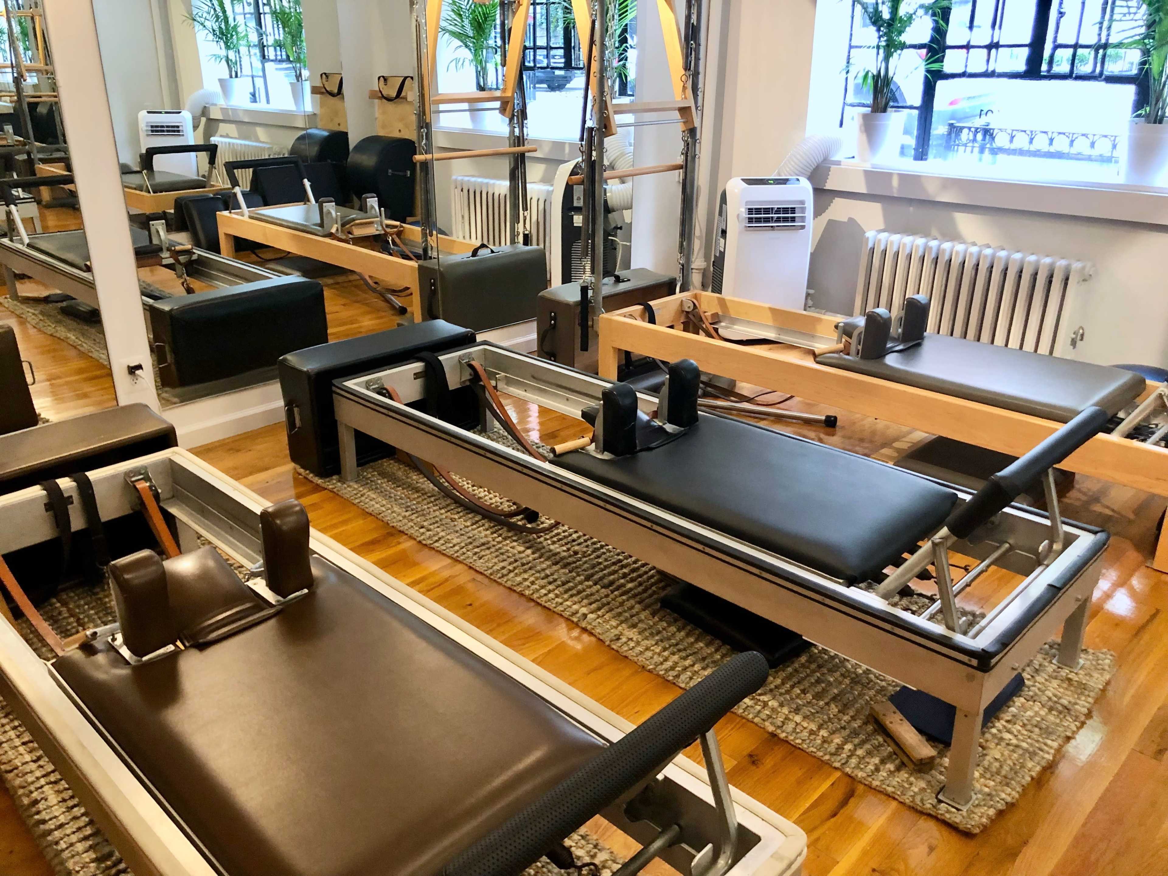 Several reformer Pilates machines arranged in a neatly organized studio with wooden flooring and plants visible through large windows.