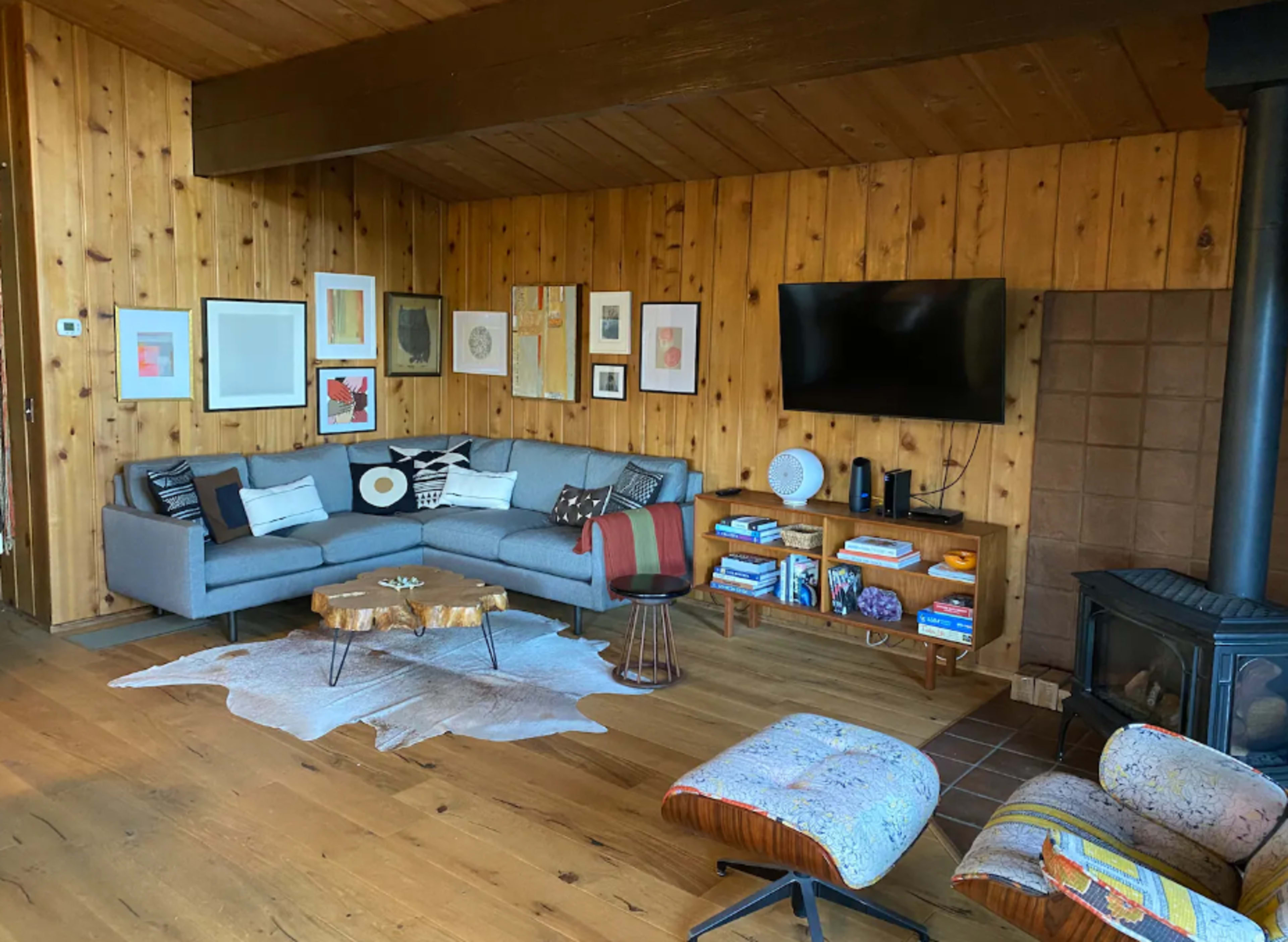 The room features a wooden-paneled interior with a gray sectional sofa, a wooden coffee table, a television mounted on the wall, and various artworks arranged on the walls.