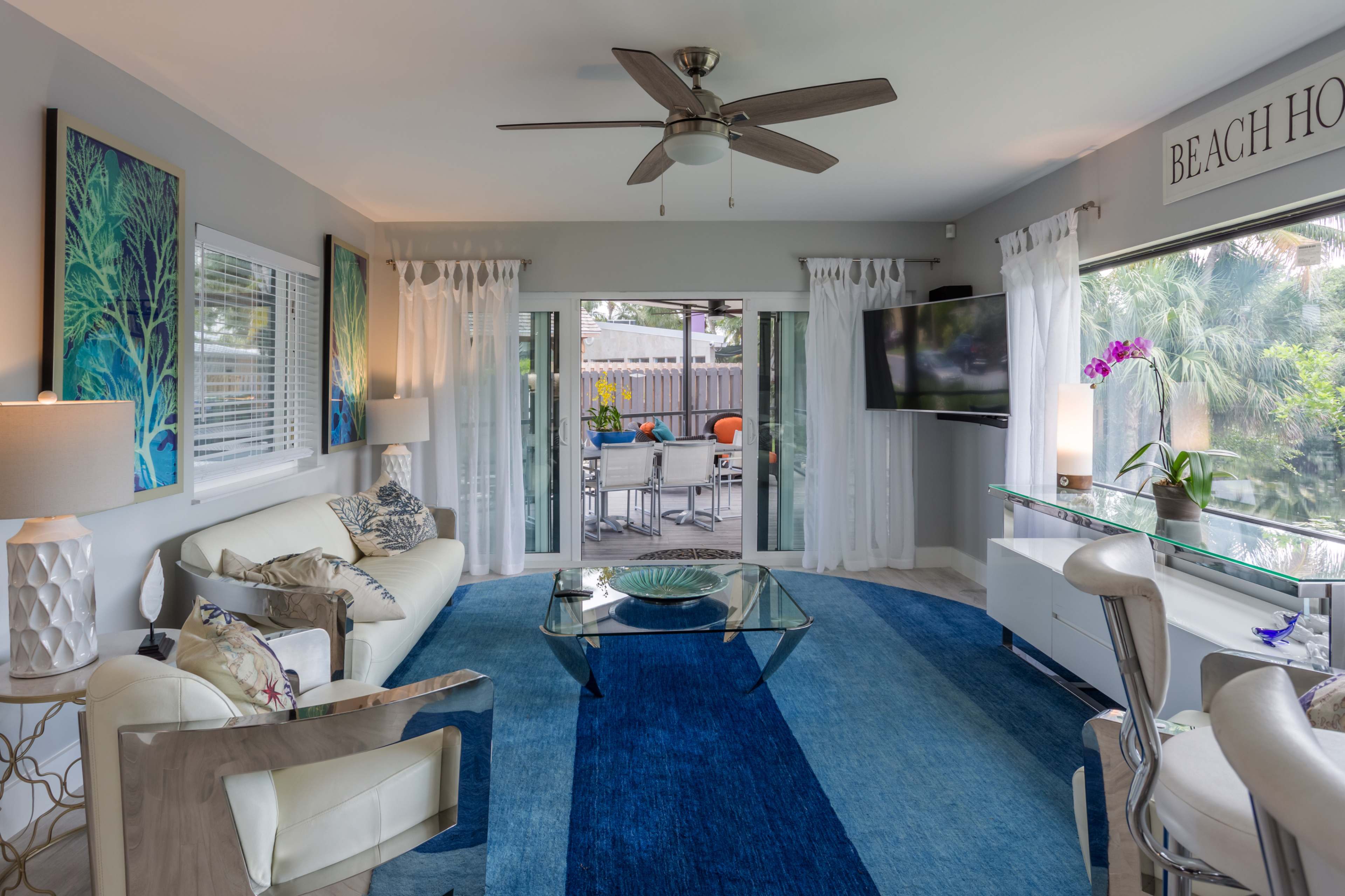 A bright living room features a white sofa, a glass coffee table, and large windows overlooking a patio area with outdoor seating.