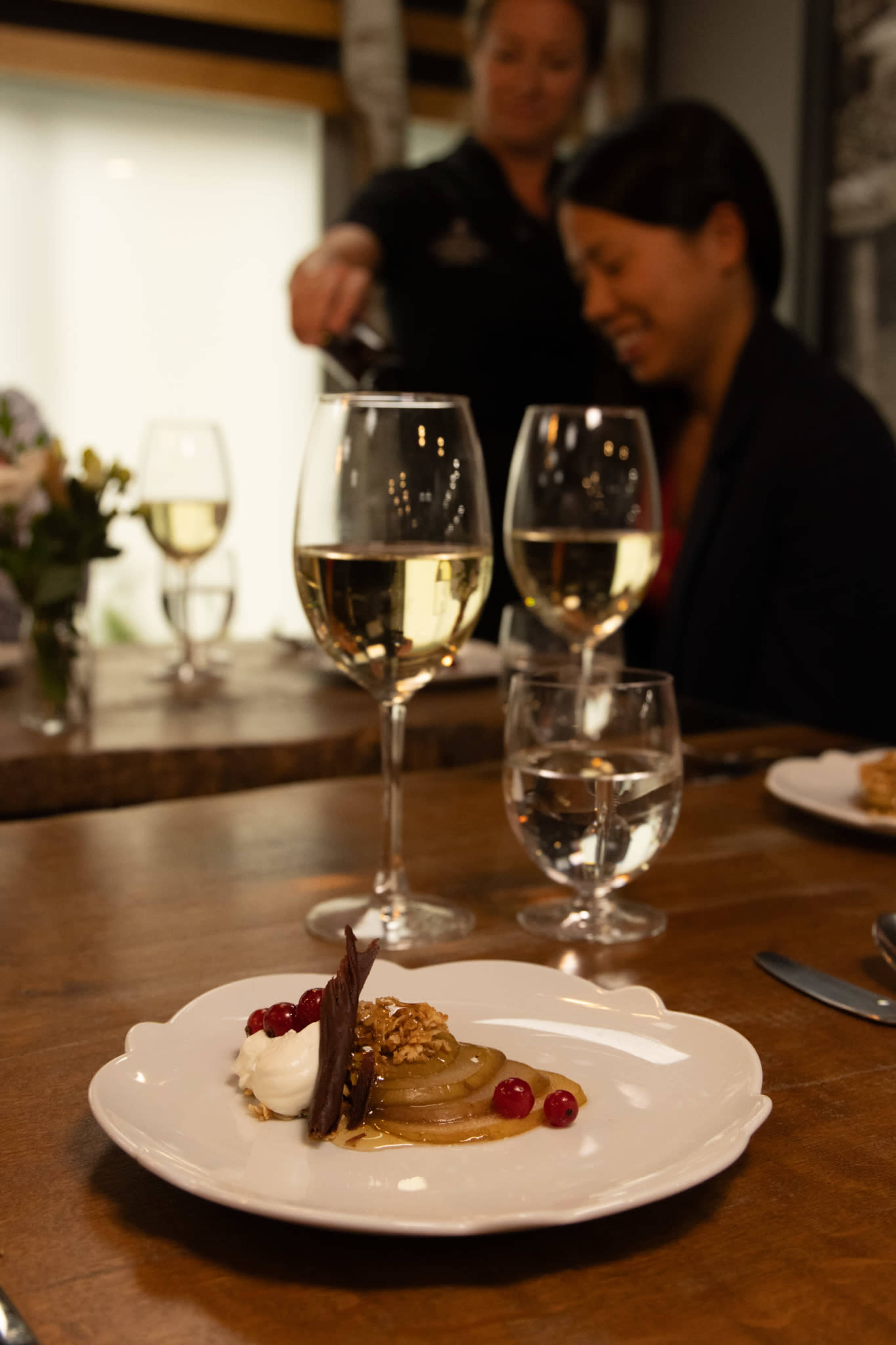 A dessert of sliced fruit, whipped cream, and chocolate garnishes is presented on a white plate, while two glasses of white wine and water sit on the wooden table beside it.