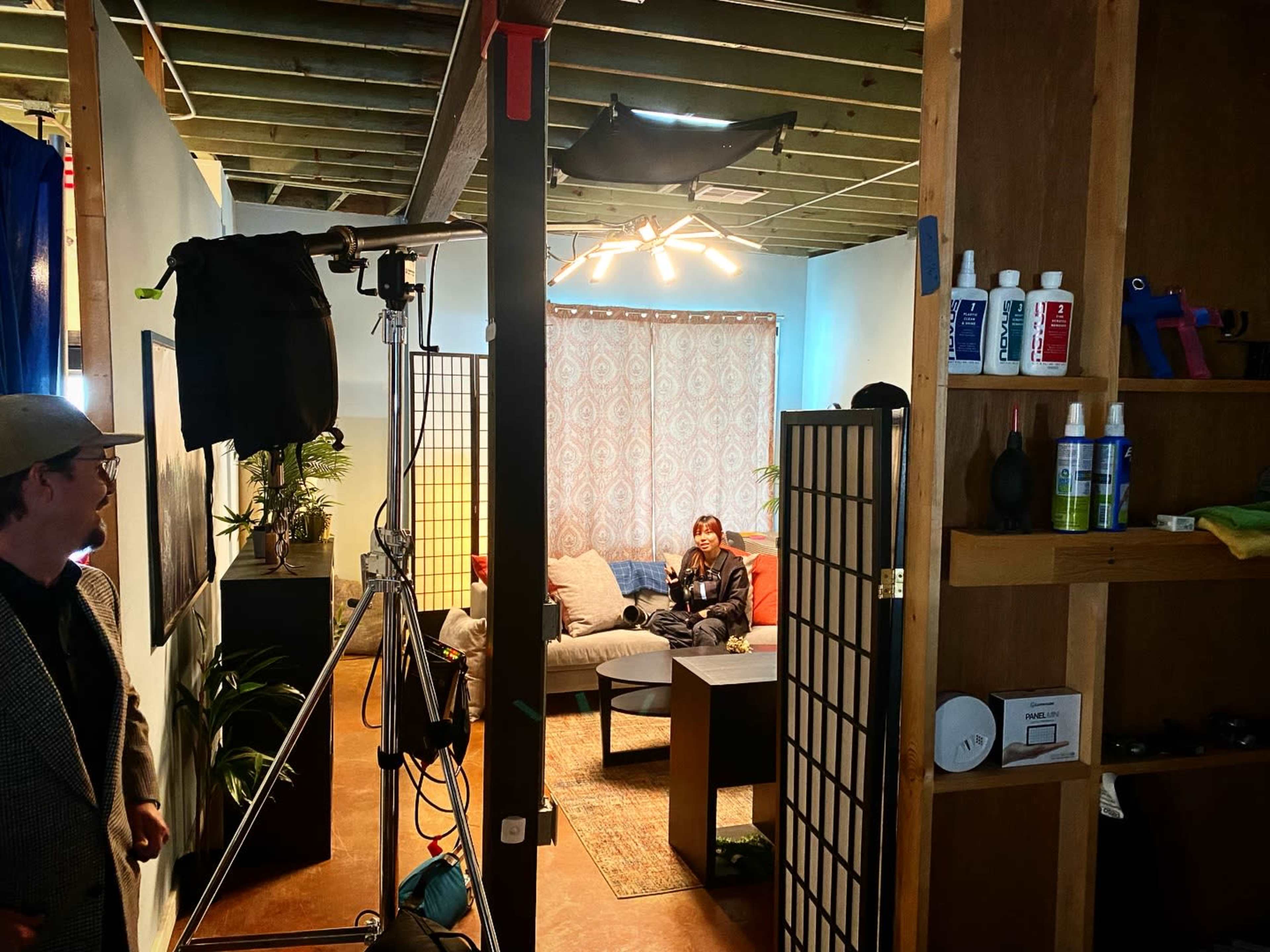 A room is set up with a couch, decorative elements, and lighting equipment, while a person sits on the couch and another stands nearby.