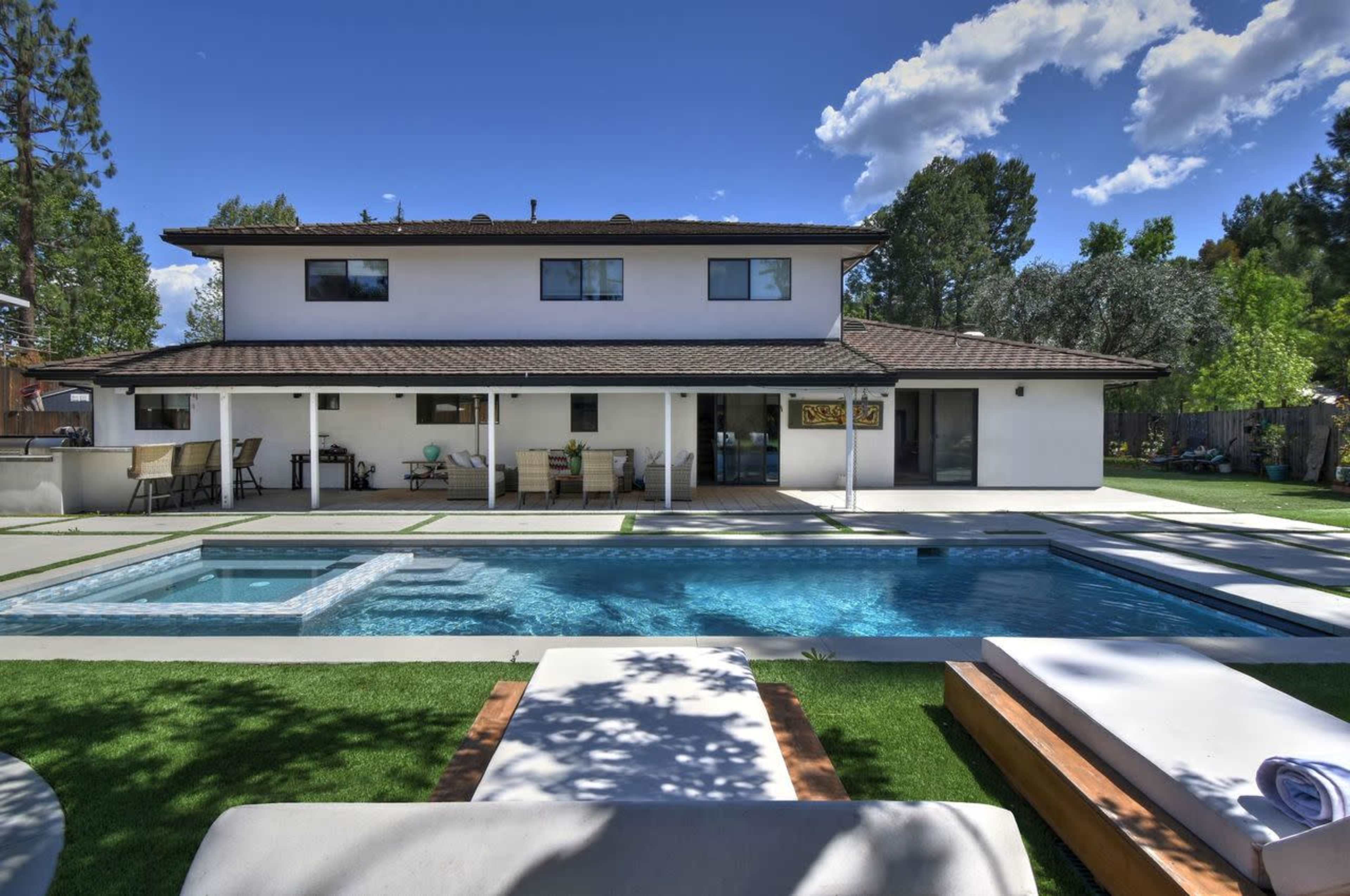 The image shows a two-story house with a landscaped backyard featuring a swimming pool, spa, and patio seating area.