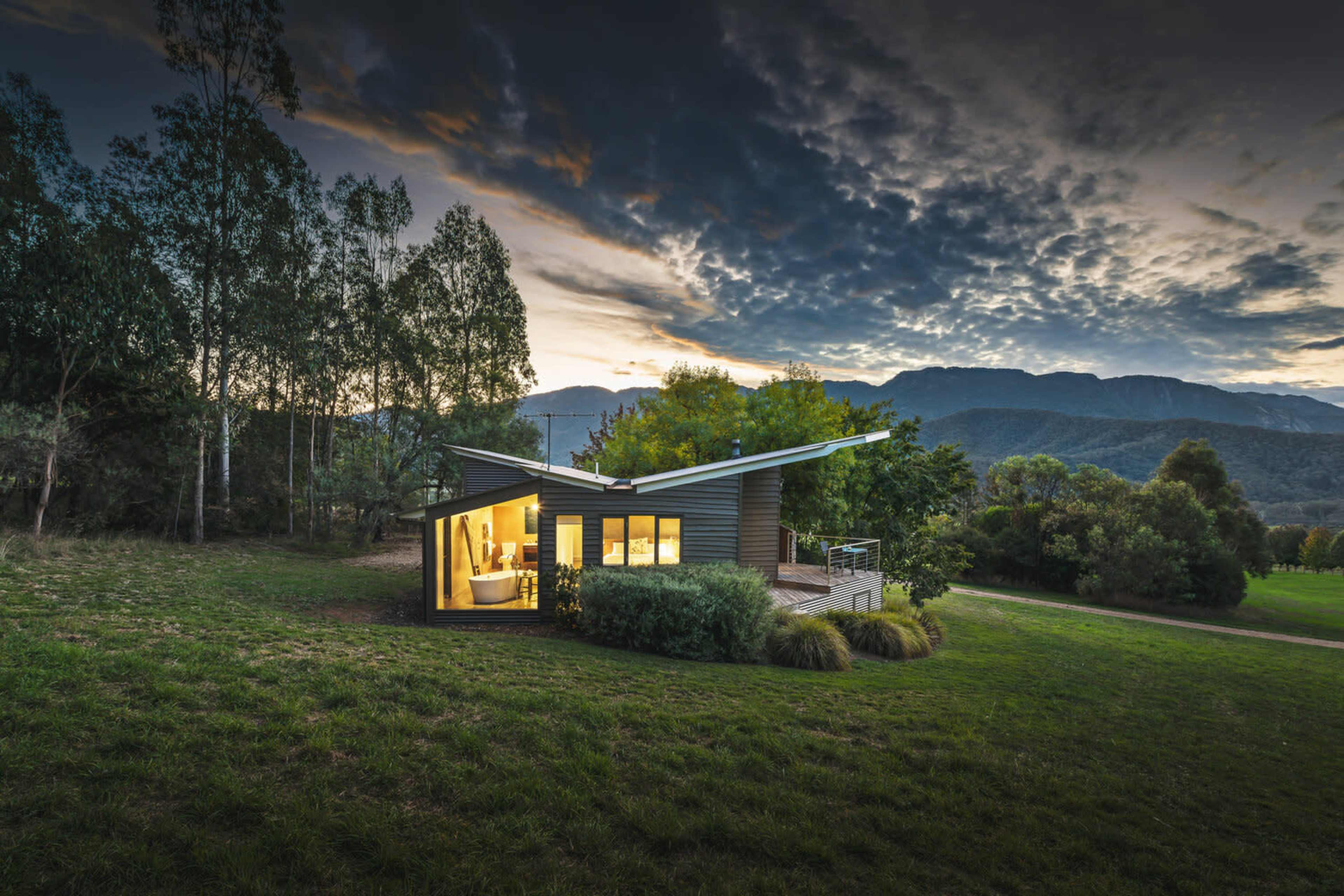 A modern, angular house with large windows is situated in a grassy area surrounded by trees, with mountains in the background under a cloudy sky.