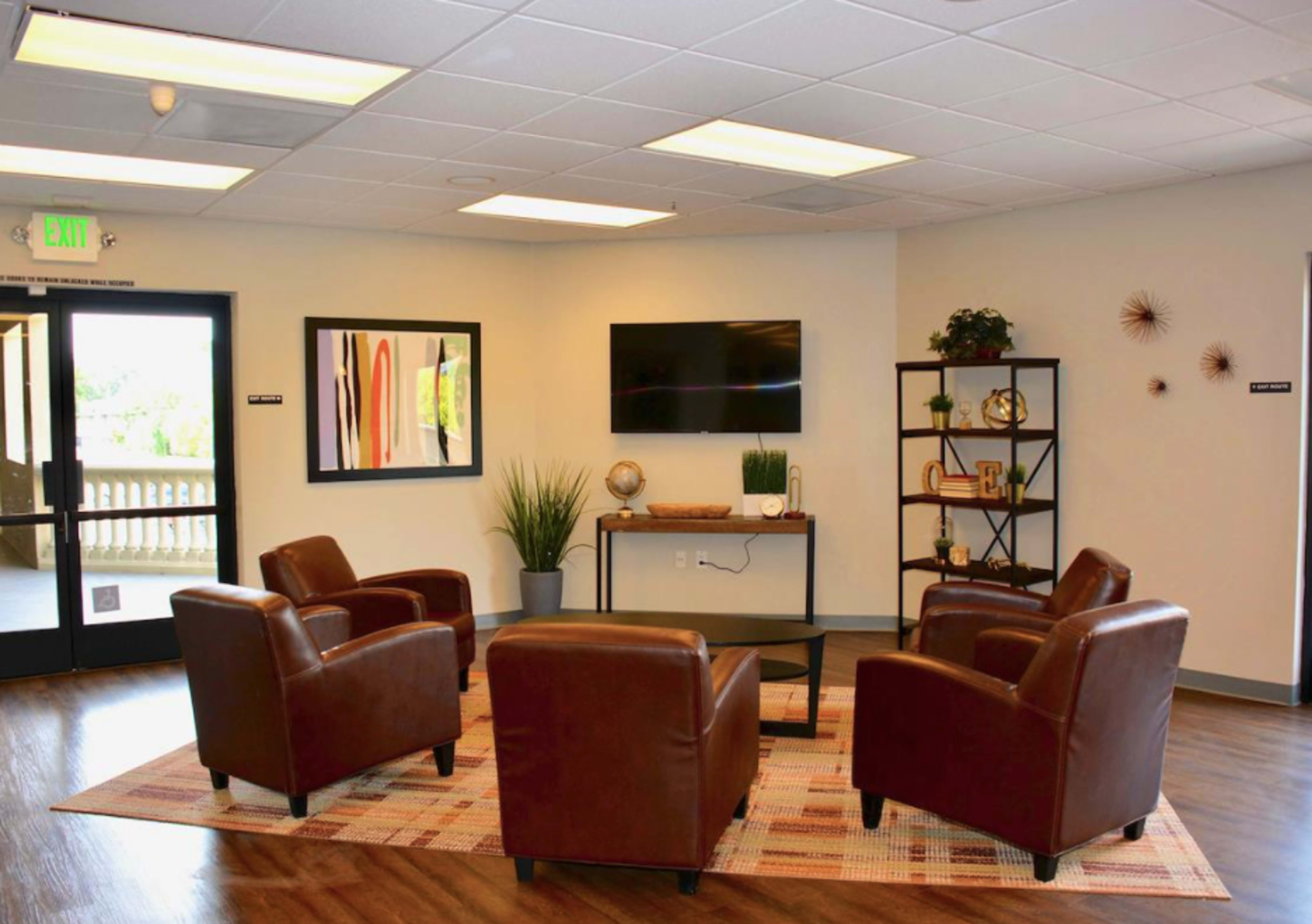 A modern lounge area with four brown chairs arranged around a coffee table, a television mounted on the wall, and decorative plants nearby.