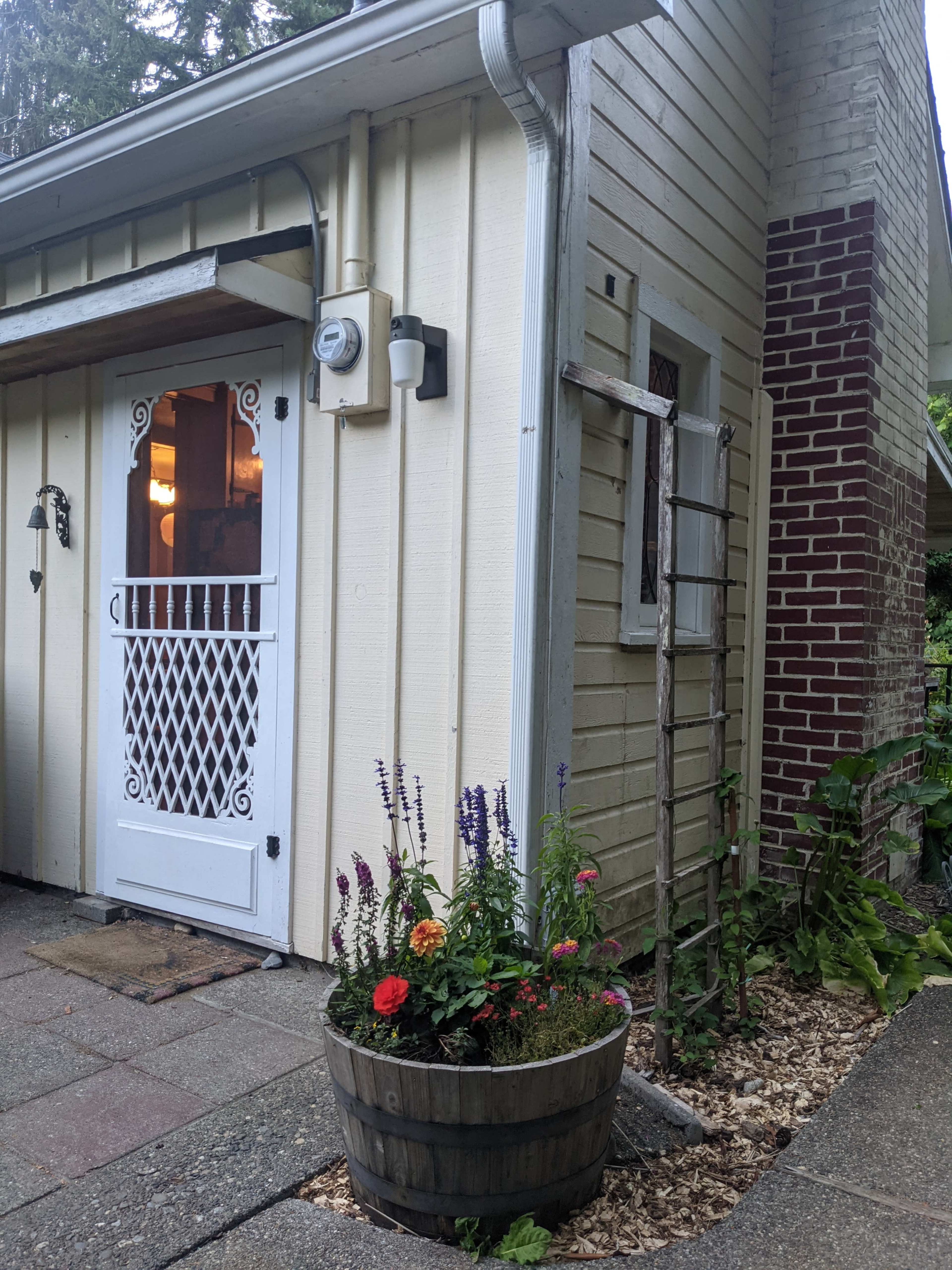 The image shows a corner of a house with a white door featuring a decorative grille, next to a flower planter filled with colorful plants and flowers.