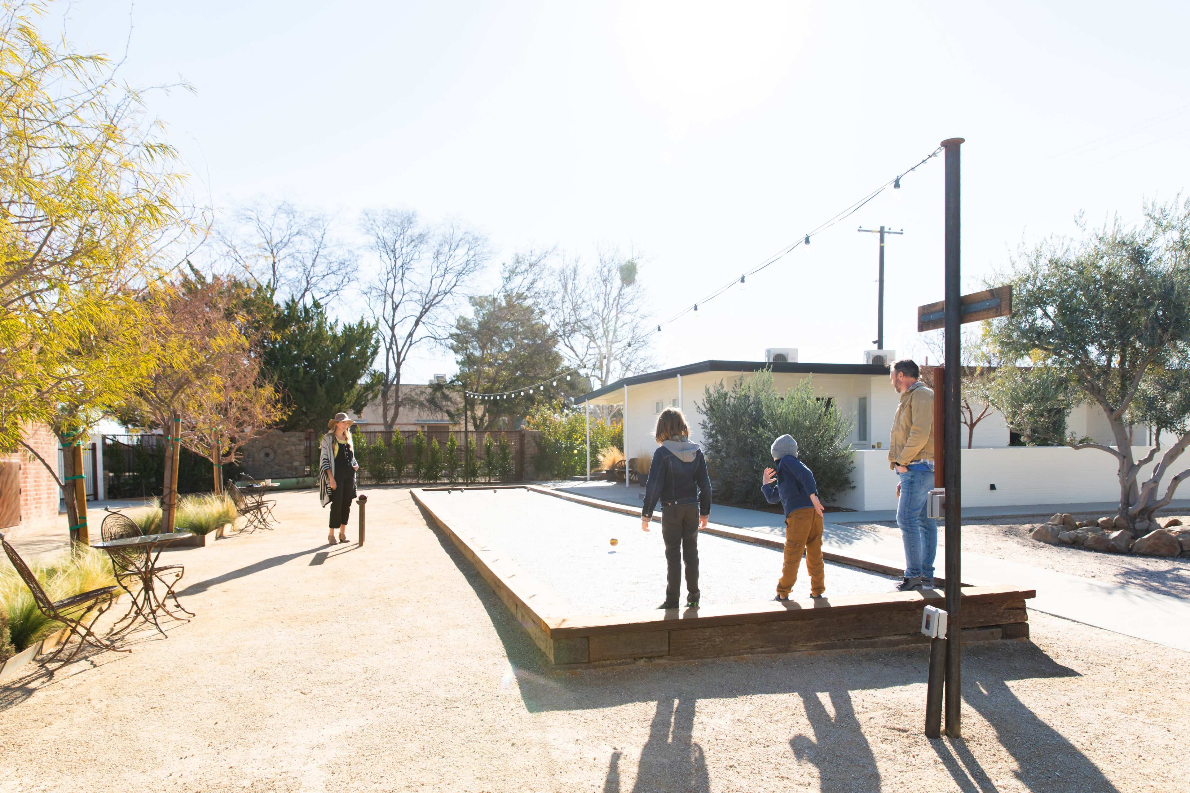 A man and two children are playing a game on a gravel court while a woman walks in the background.