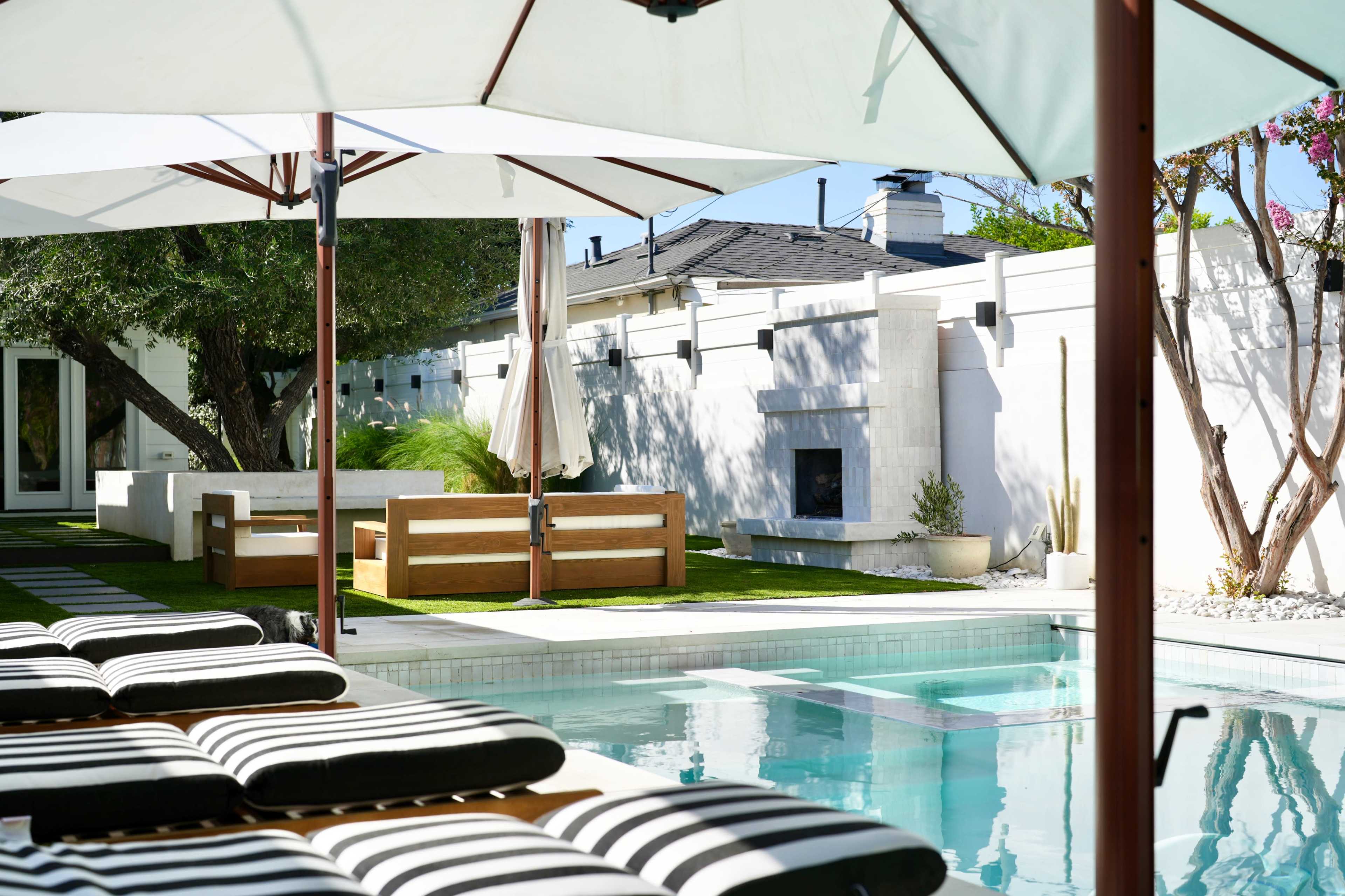 A crystal-clear swimming pool reflects the sun, surrounded by lounge chairs and a landscaped yard with a modern seating area and an outdoor fireplace.