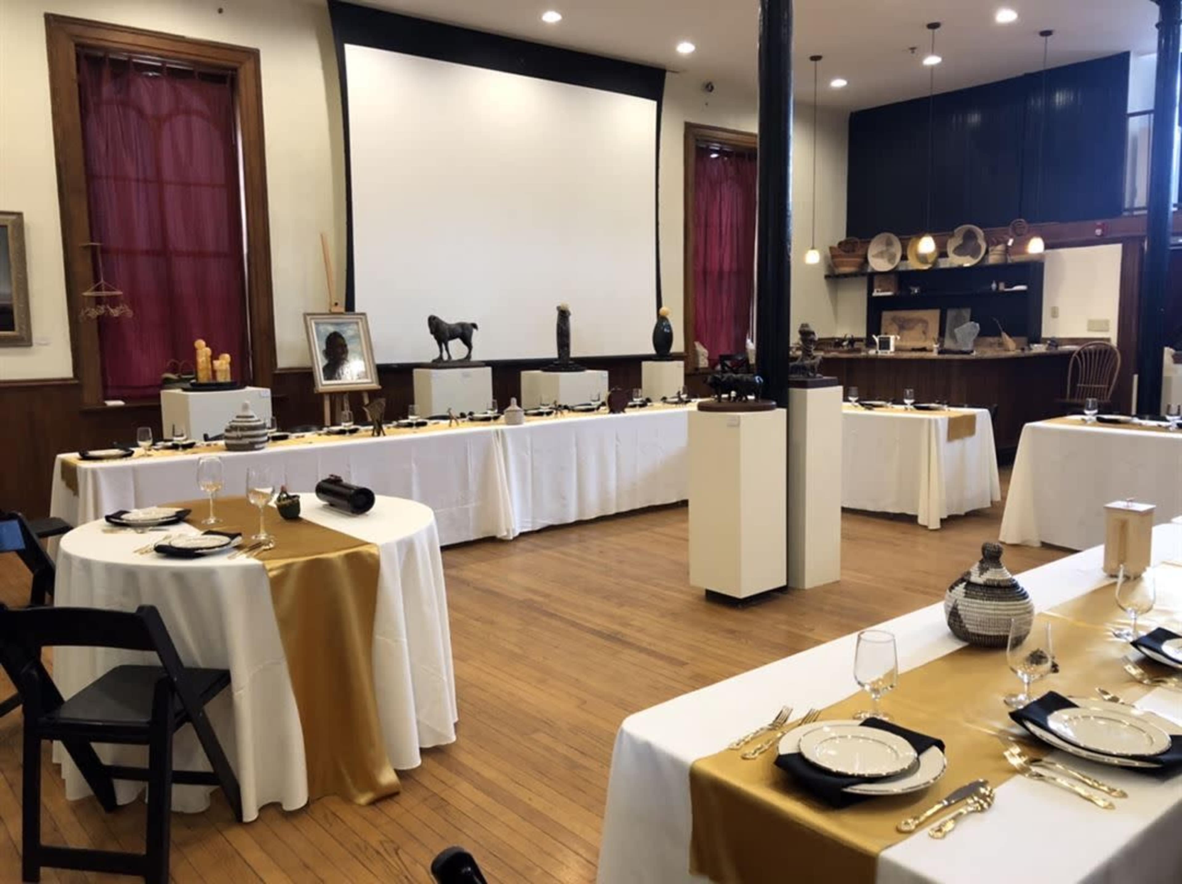 The image shows a spacious dining area with several elegantly set tables covered in white and gold tablecloths, surrounded by art displays.