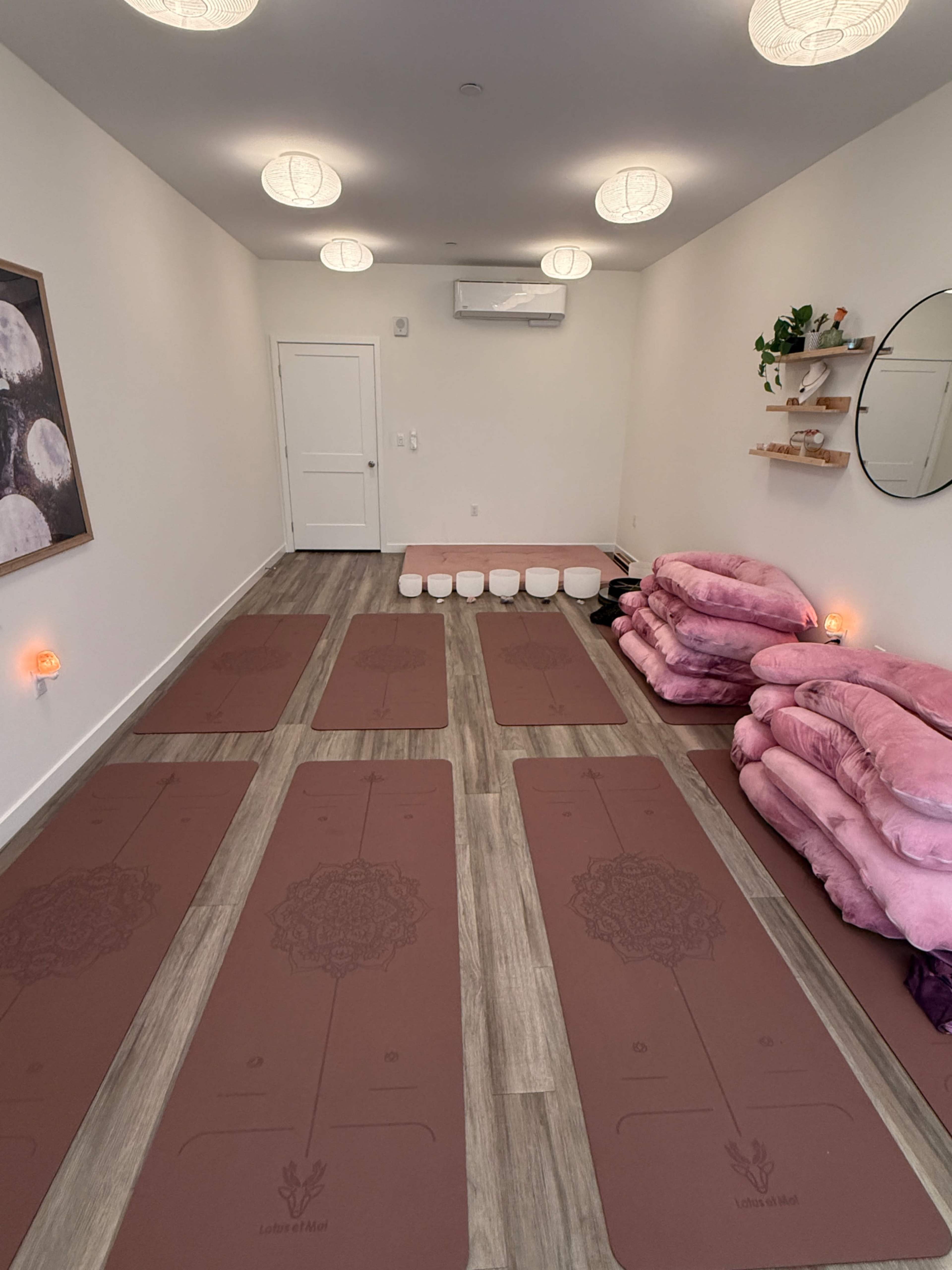 The image shows a serene yoga studio with neatly arranged yoga mats, a row of white bolsters, and soft pink blankets on a shelf, all set against light-colored walls.