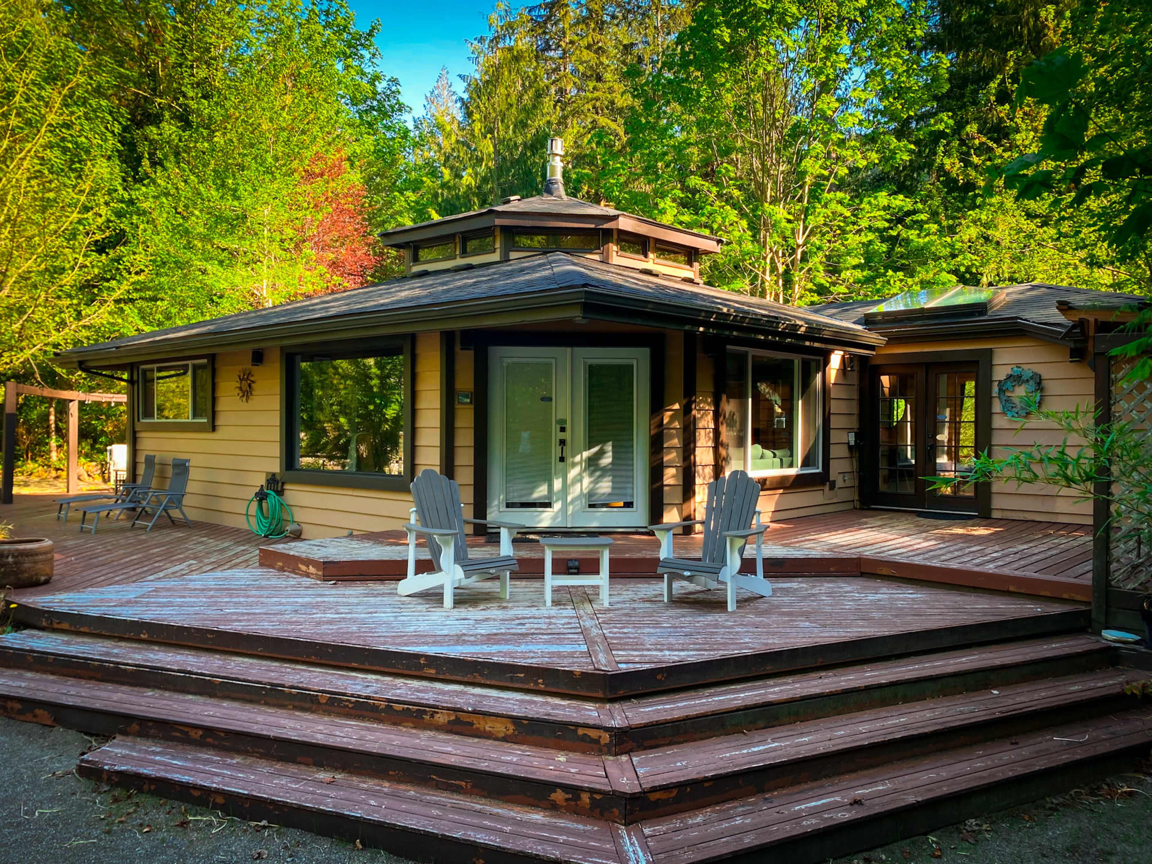 A modern, single-story house surrounded by trees, with a wooden deck featuring a seating area in front.