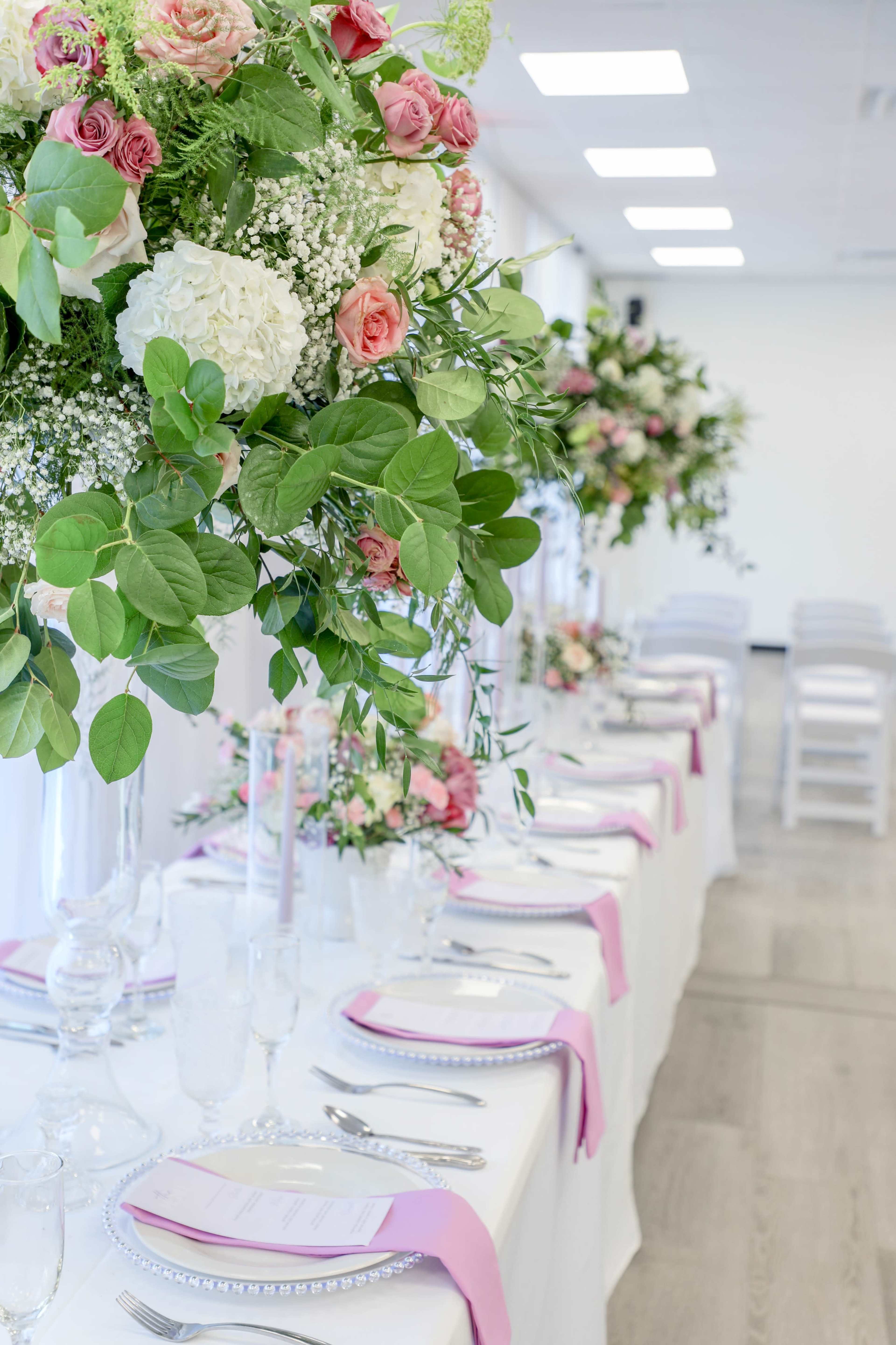 A long banquet table is elegantly set with silverware, pink napkins, and floral centerpieces, surrounded by white chairs in a bright indoor space.