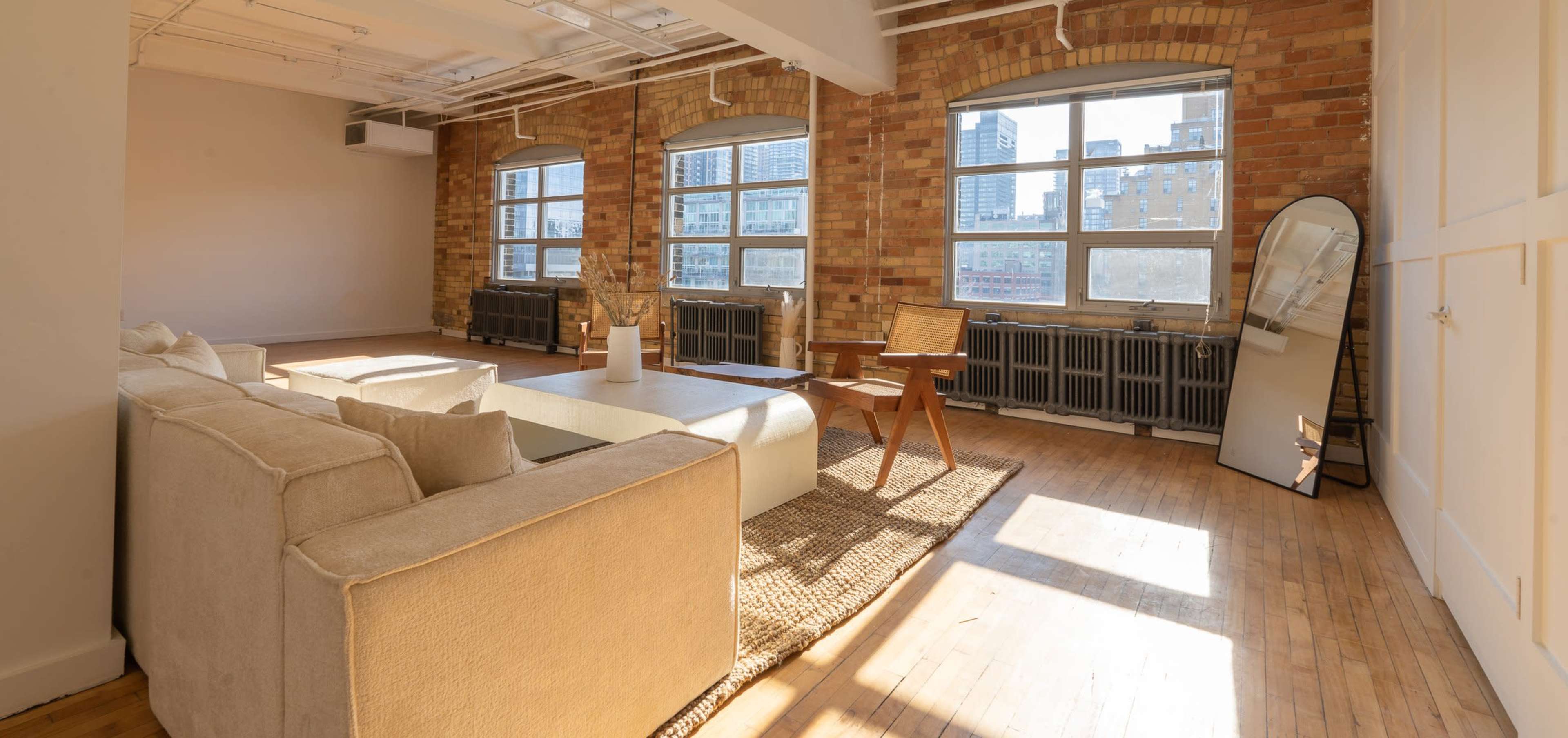 A spacious, sunlit living room features large windows, a beige sectional sofa, a wooden chair, and a large mirror against a brick wall.