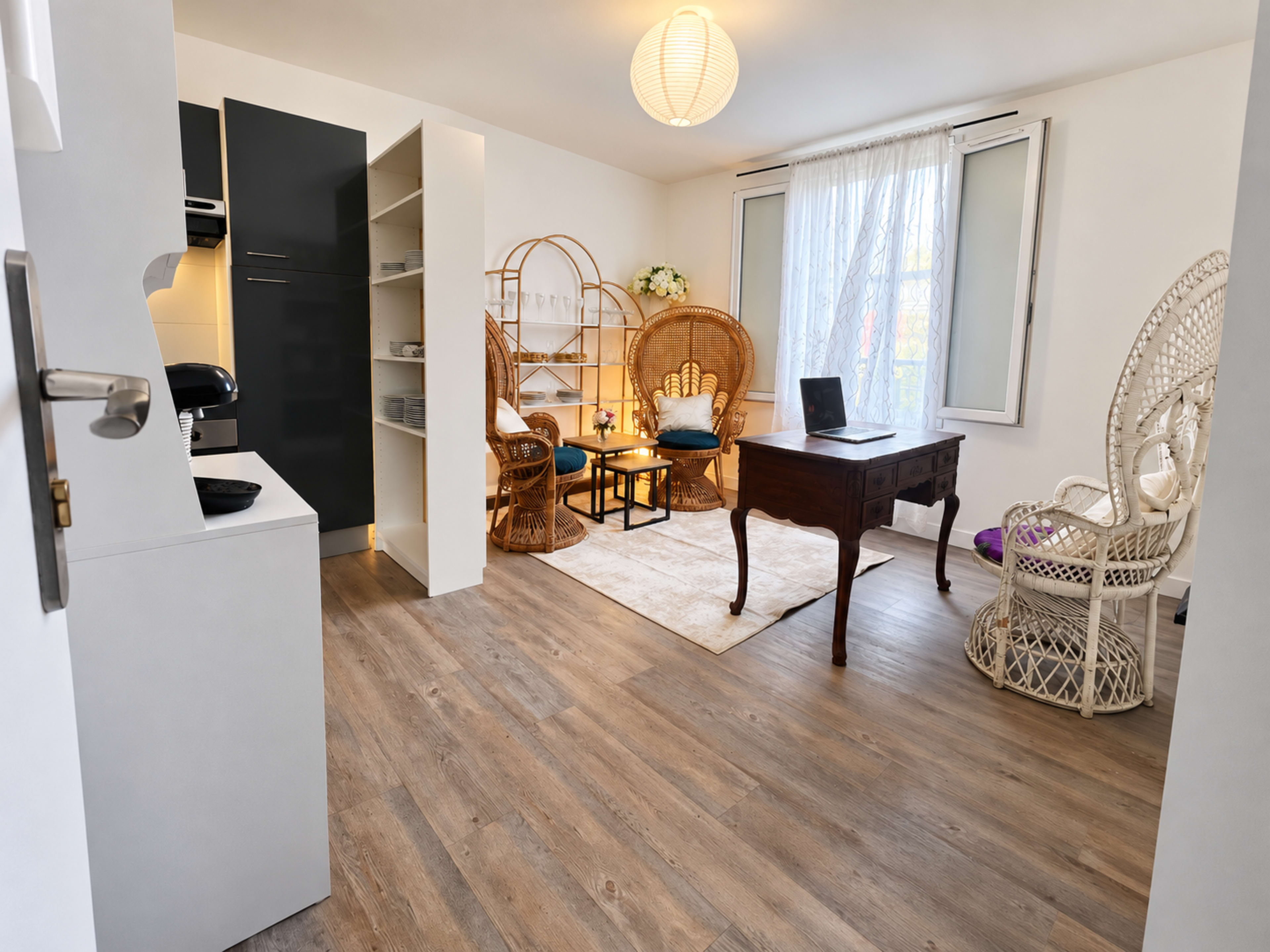 The image shows a tidy, bright room featuring a small kitchen area, a wooden desk, and two wicker seating arrangements beside a large window.