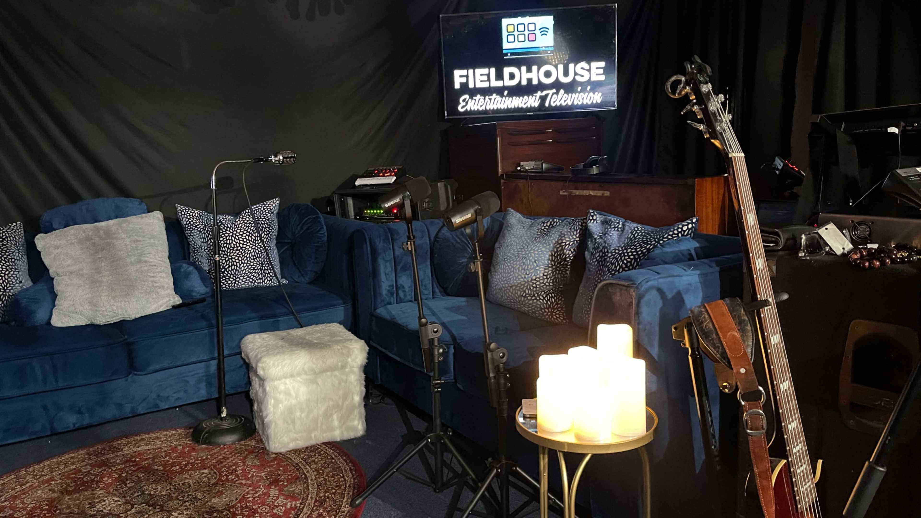 A cozy studio setup with blue couches, a guitar, microphones, and a sign that reads "FIELDHOUSE Entertainment Television."