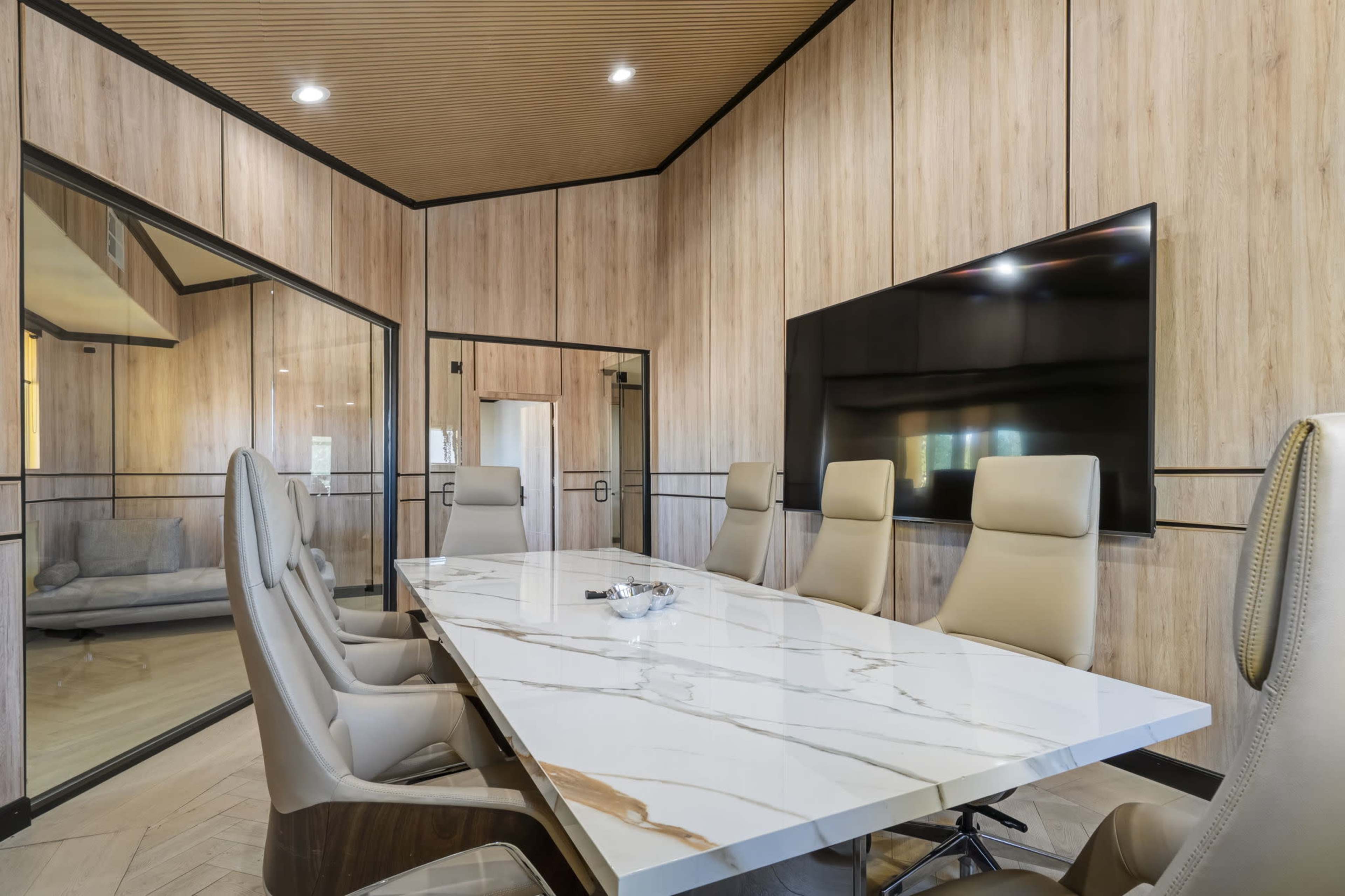 The image shows a modern conference room featuring a large marble table surrounded by high-backed chairs, with a wall-mounted television and wood-paneled walls.