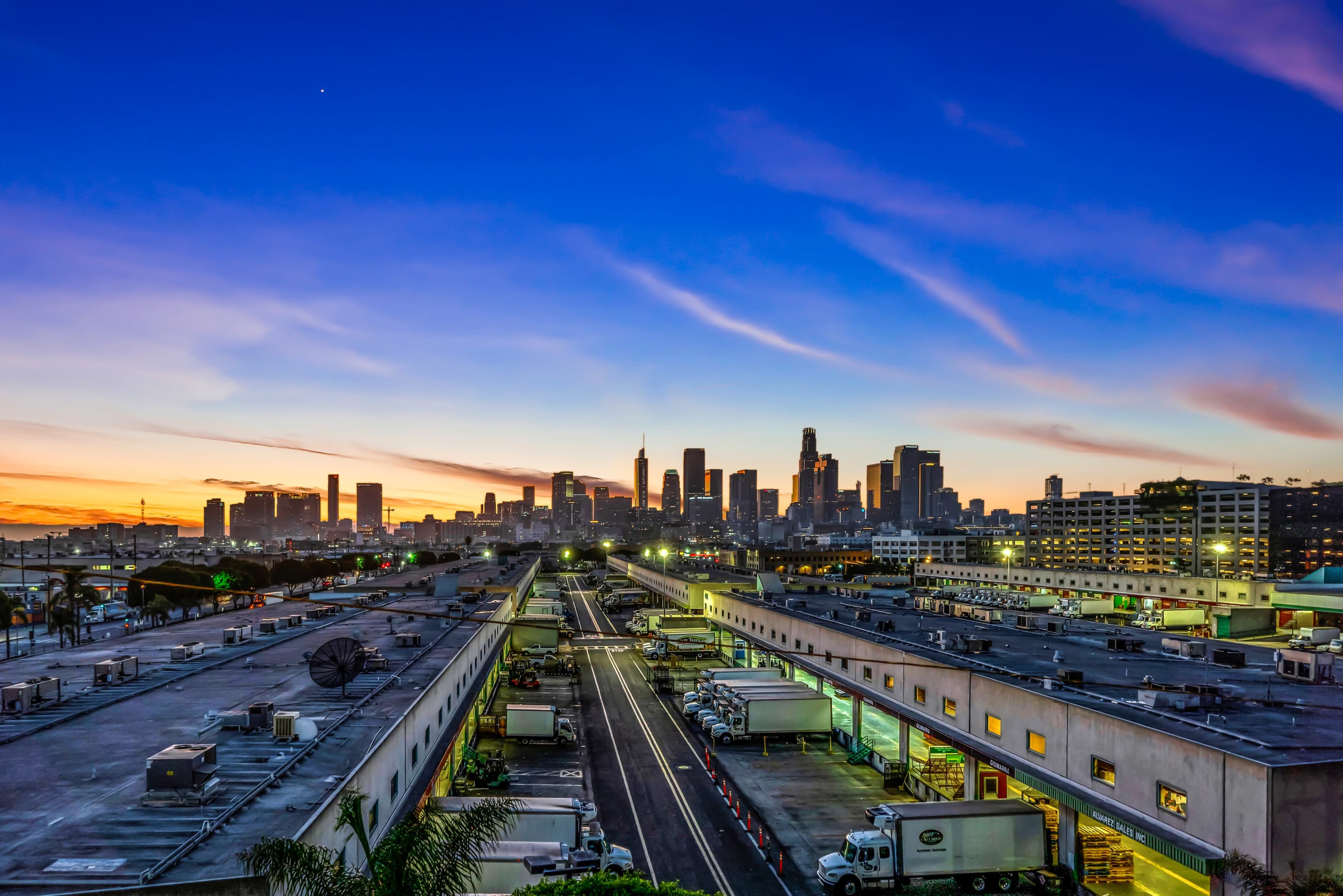 A city skyline at sunset, with trucks lined up in a distribution facility in the foreground.
