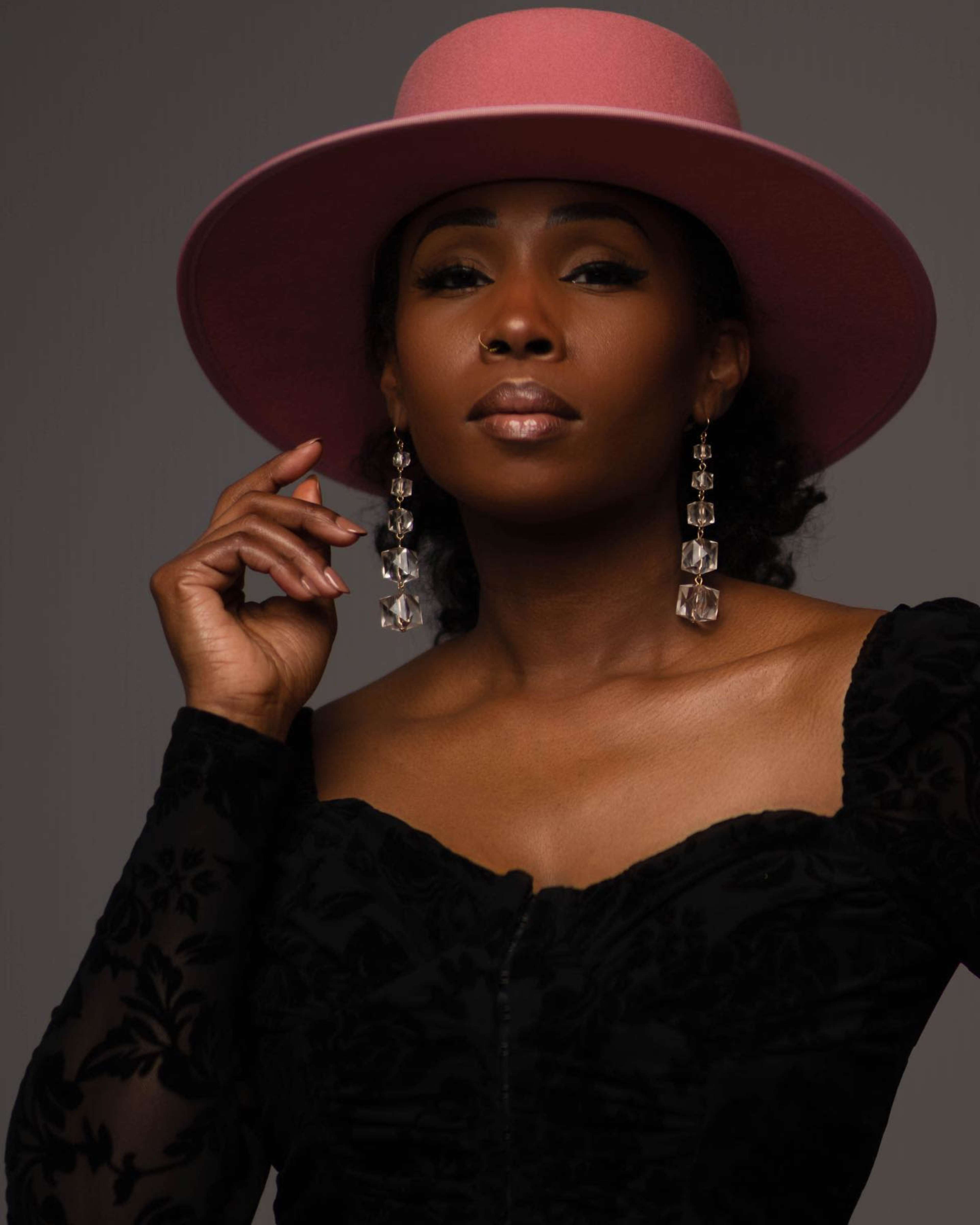 A woman wearing a pink wide-brimmed hat and large earrings poses with a hand near her face against a neutral background.
