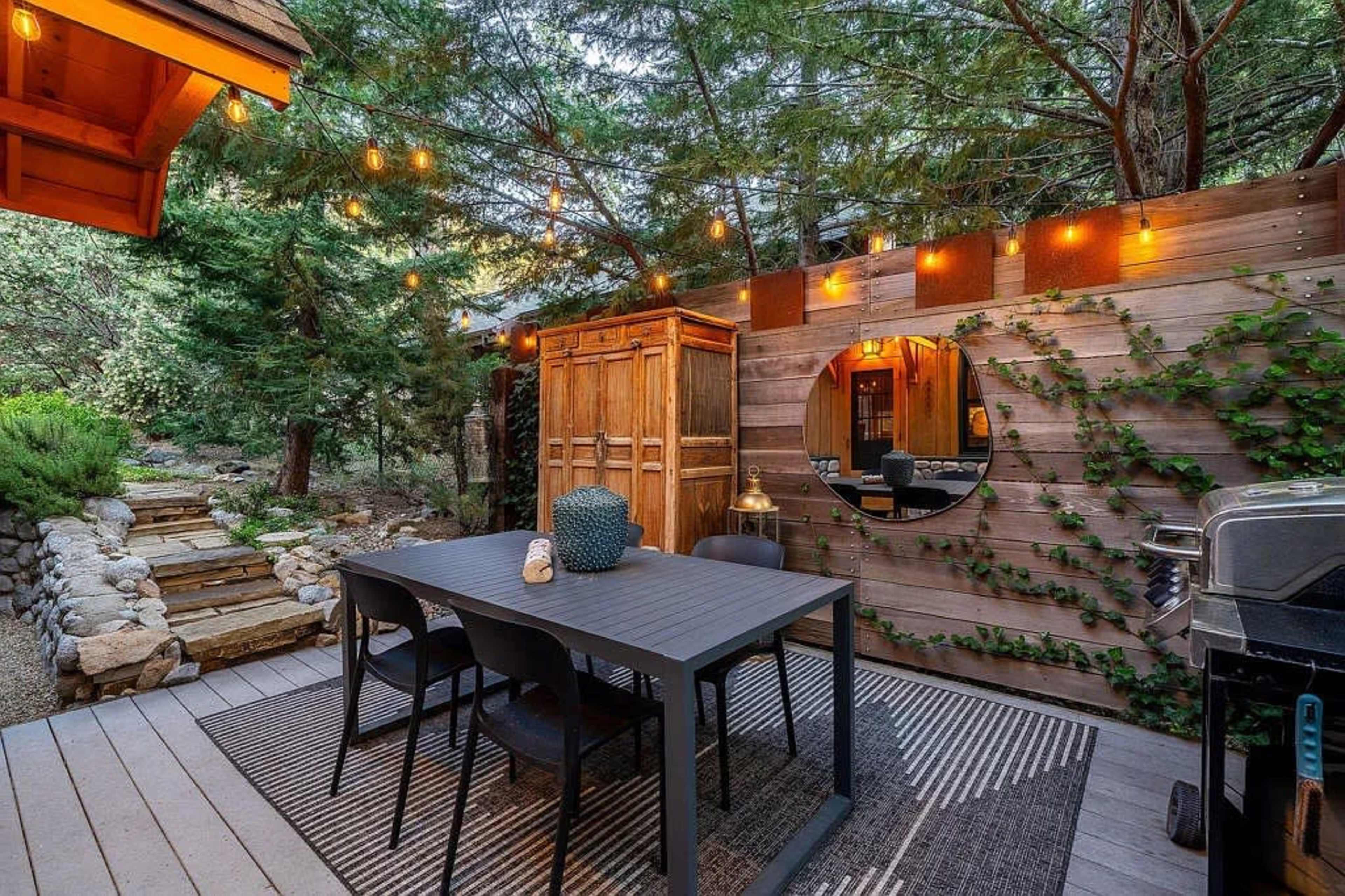 The image shows an outdoor dining area with a table, chairs, and a grill, surrounded by trees and decorative lighting.