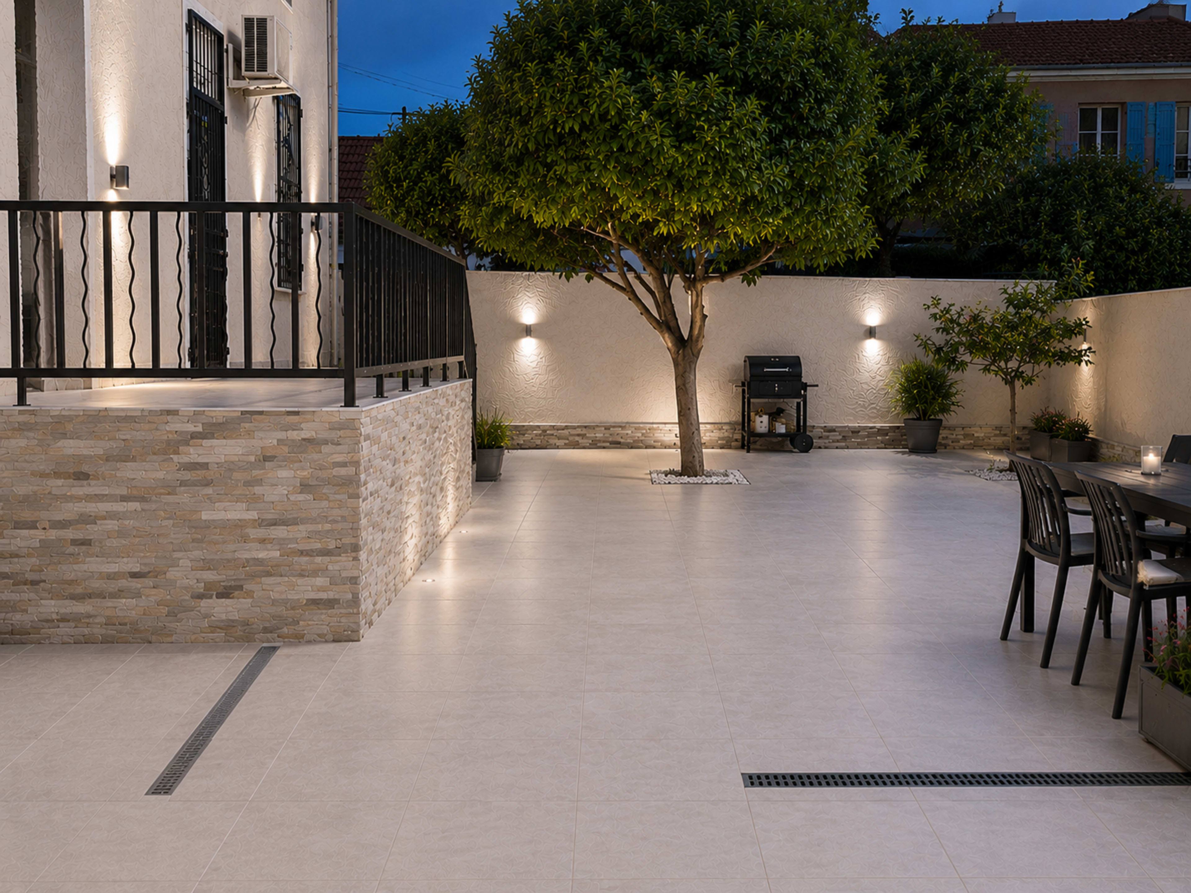 The image shows a neatly arranged outdoor patio with a stone wall, a dining table, a grill area, and a well-maintained tree, illuminated by wall lights.