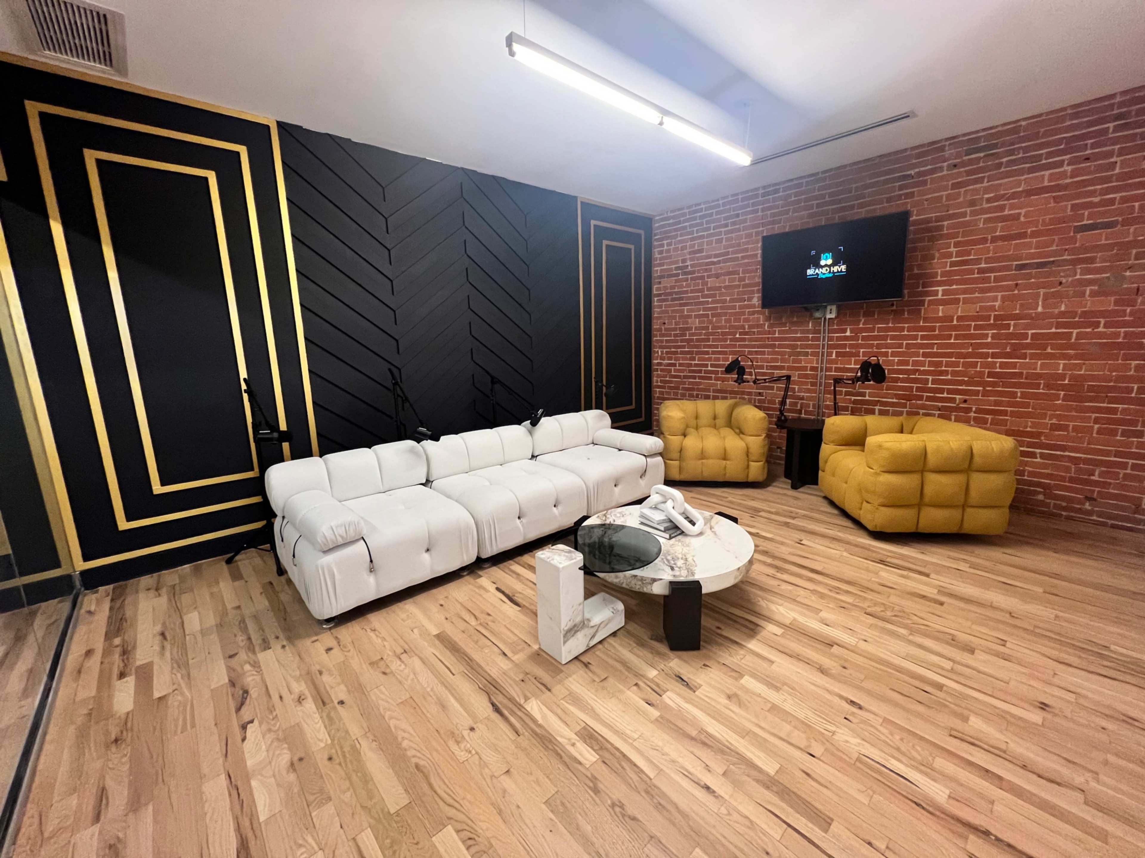 The scene features a modern lounge area with a white sectional sofa, two yellow armchairs, a round coffee table, and a brick wall accentuated by black and gold decor.