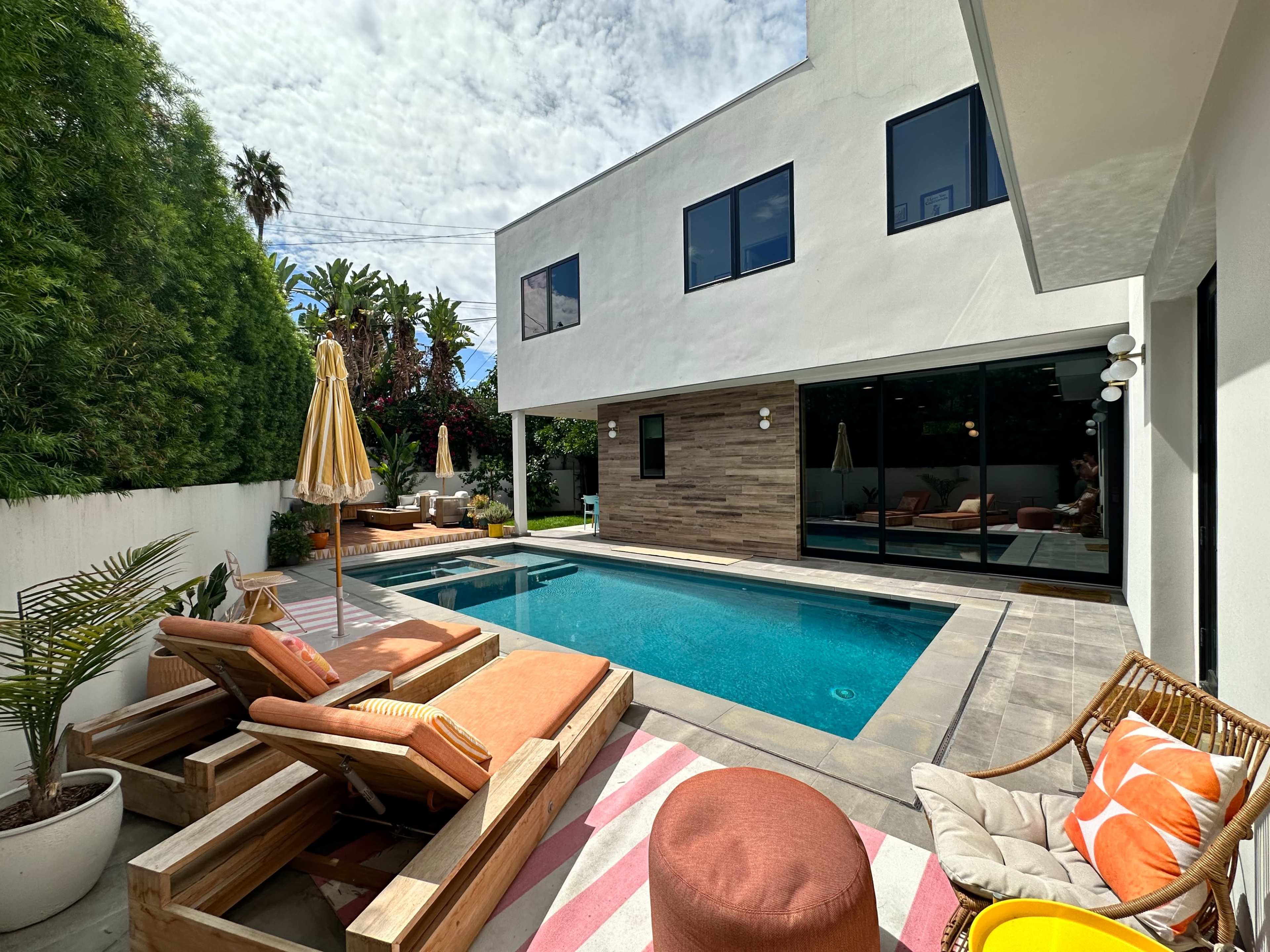 A modern backyard features a swimming pool, lounge chairs, and a patio area with seating, surrounded by lush greenery.