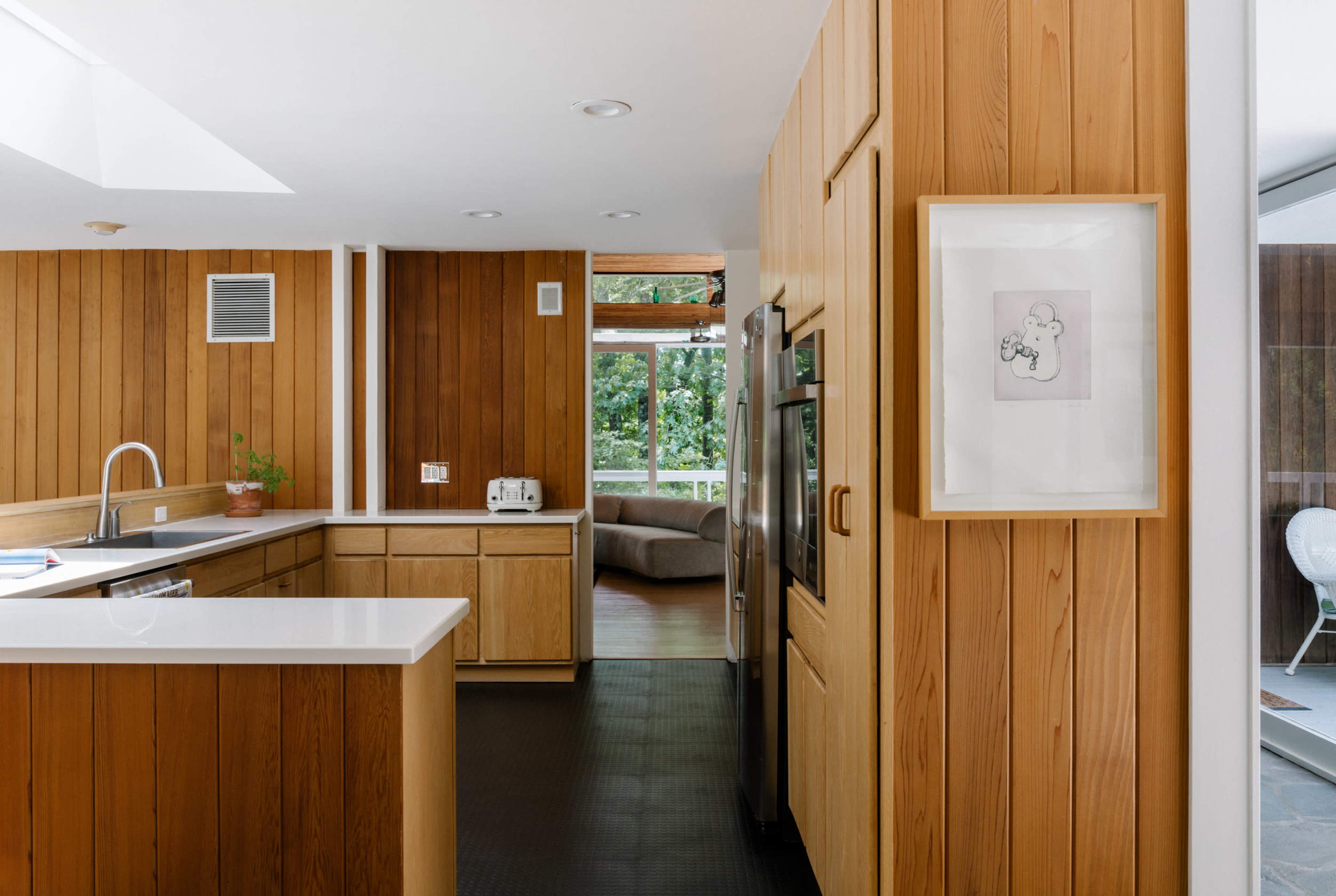 The kitchen features wooden paneling, a white countertop, and stainless steel appliances, with a view of a living area through a large window.