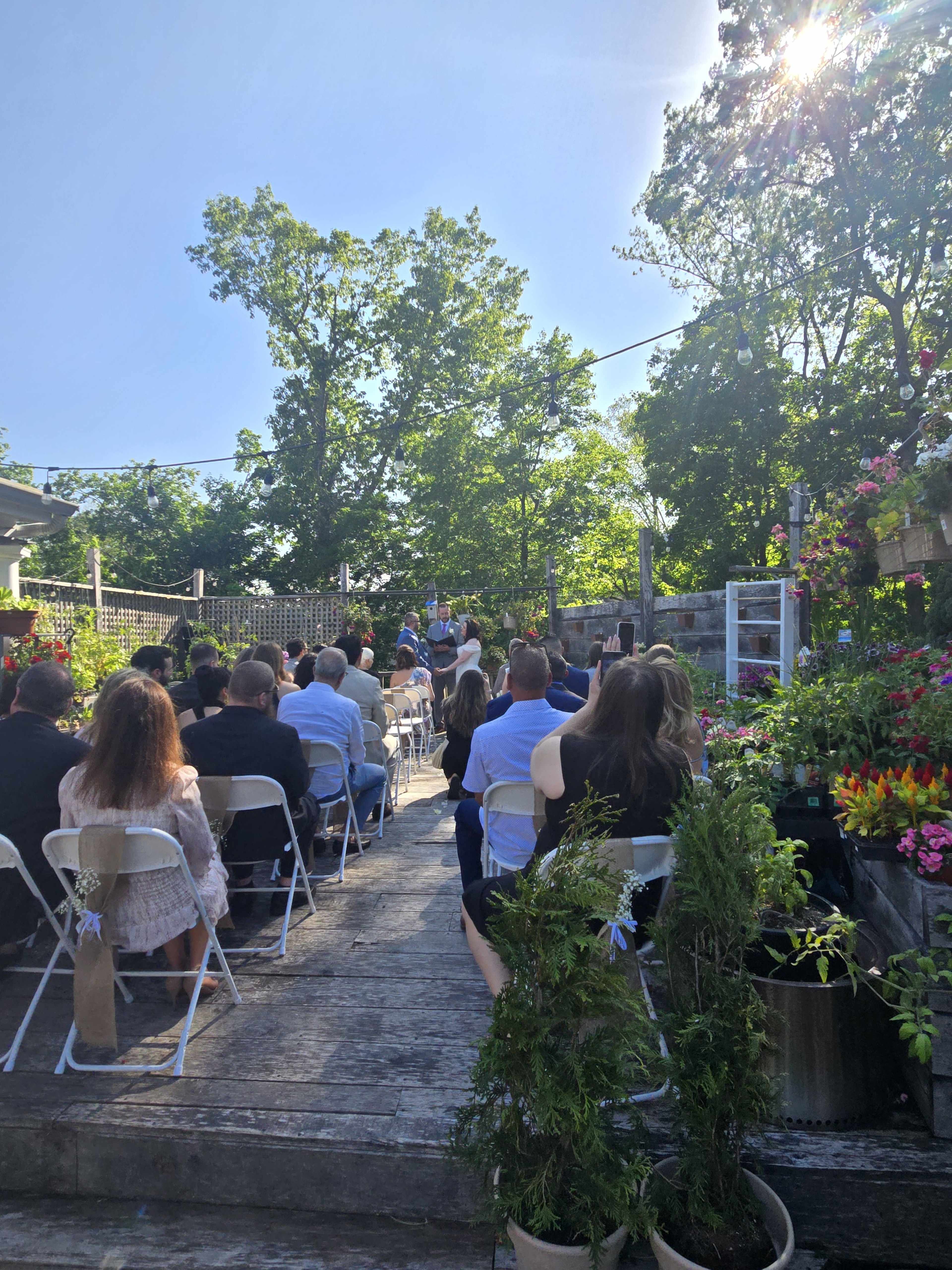 A wedding ceremony takes place outdoors with guests seated on chairs arranged in rows among greenery and flowers.
