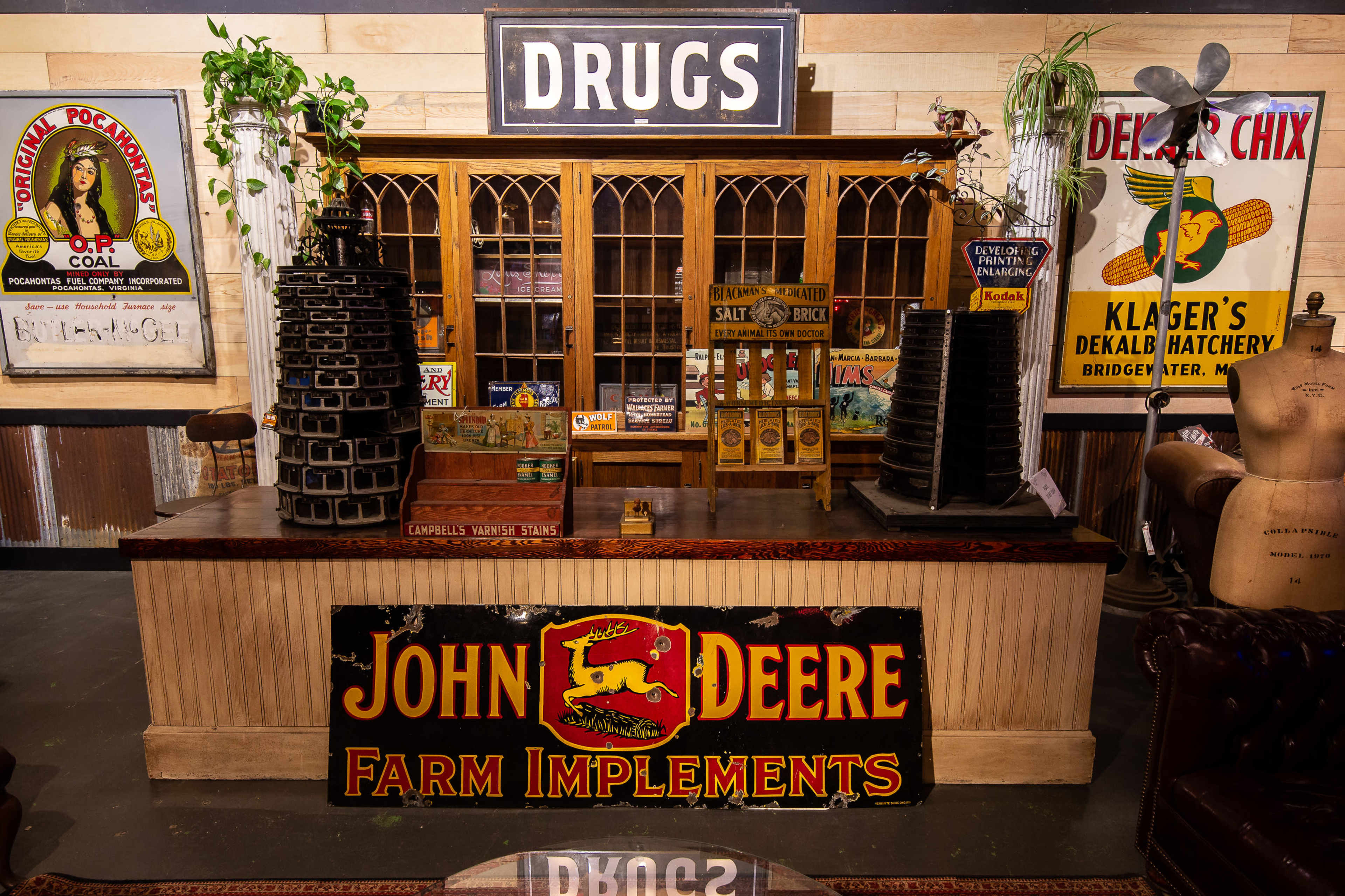 The image showcases a vintage pharmacy display counter with a prominent "DRUGS" sign and a "JOHN DEERE" Farm Implements sign in front.