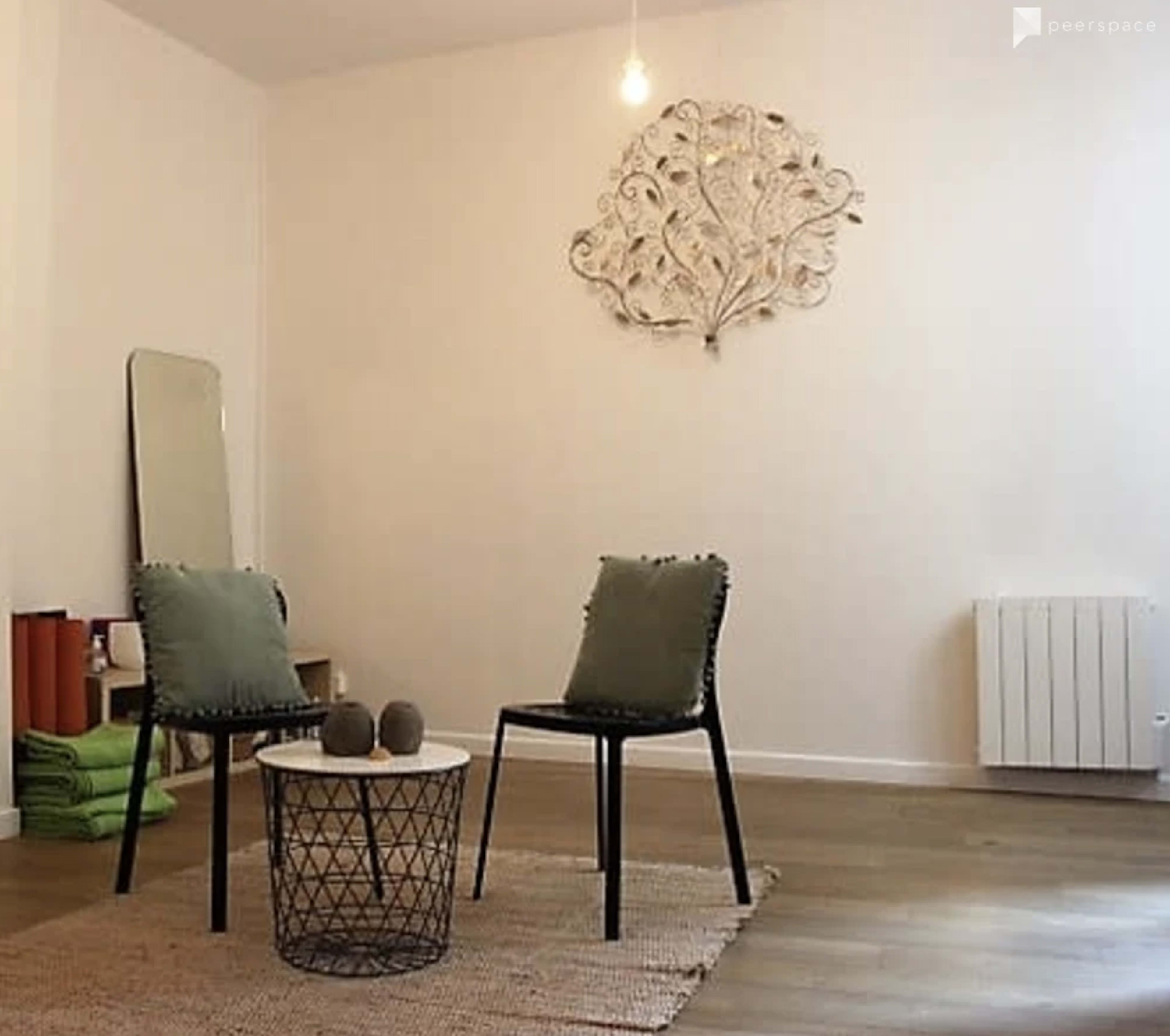 A minimalistic interior space with two chairs, a round table, and a decorative wall piece, featuring a mirror, a radiator, and neatly arranged green towels in the background.