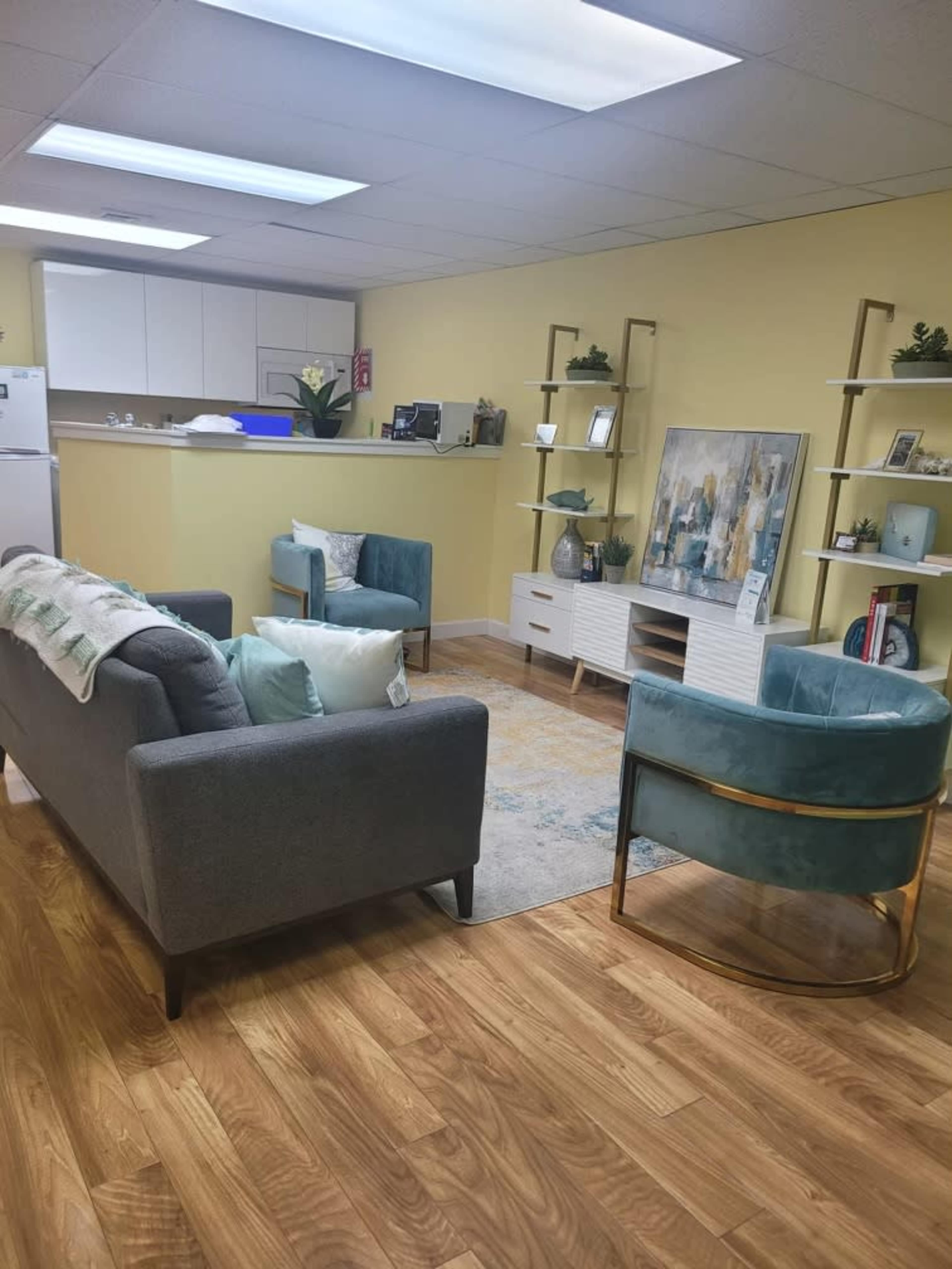 A modern reception area with two seating areas, a coffee table, a wall-mounted shelf, and a kitchenette in the background.
