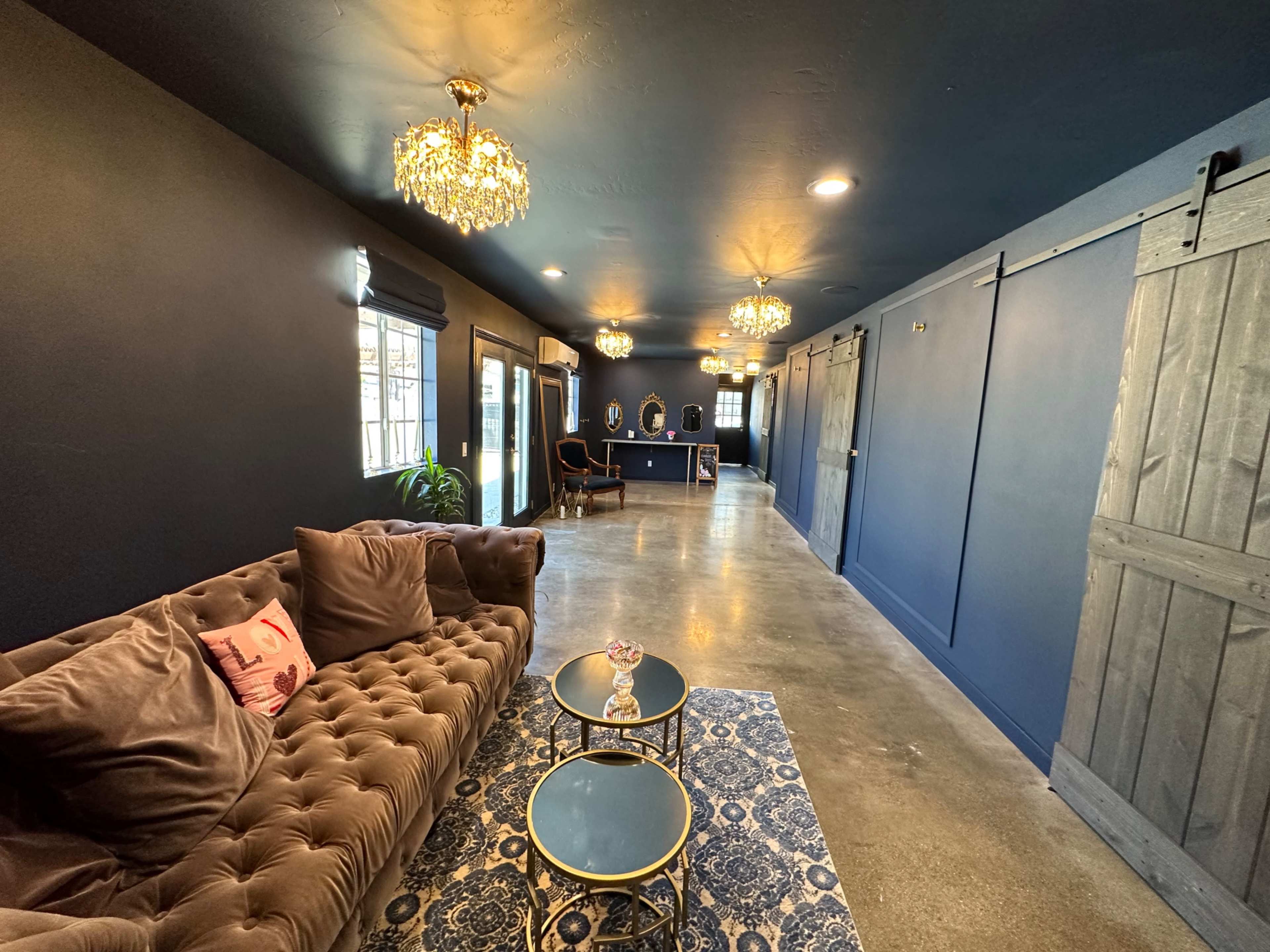 A long, narrow lounge area featuring a tufted brown sofa, two small coffee tables, and a dark blue wall with elegant chandeliers and sliding barn doors.