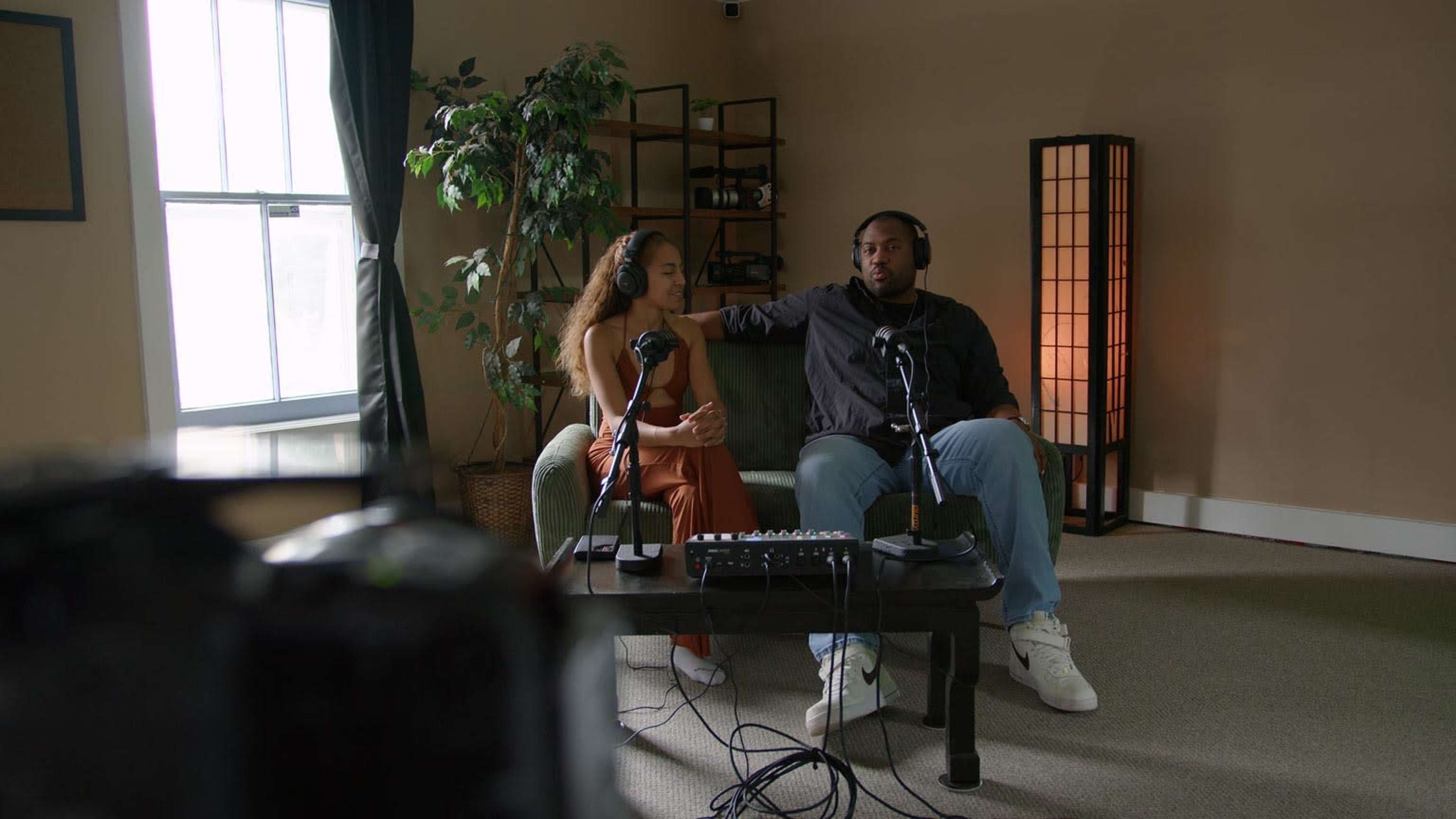 A man and a woman sit on a green couch in a well-lit room, wearing headphones and microphones, with audio equipment on a table between them.