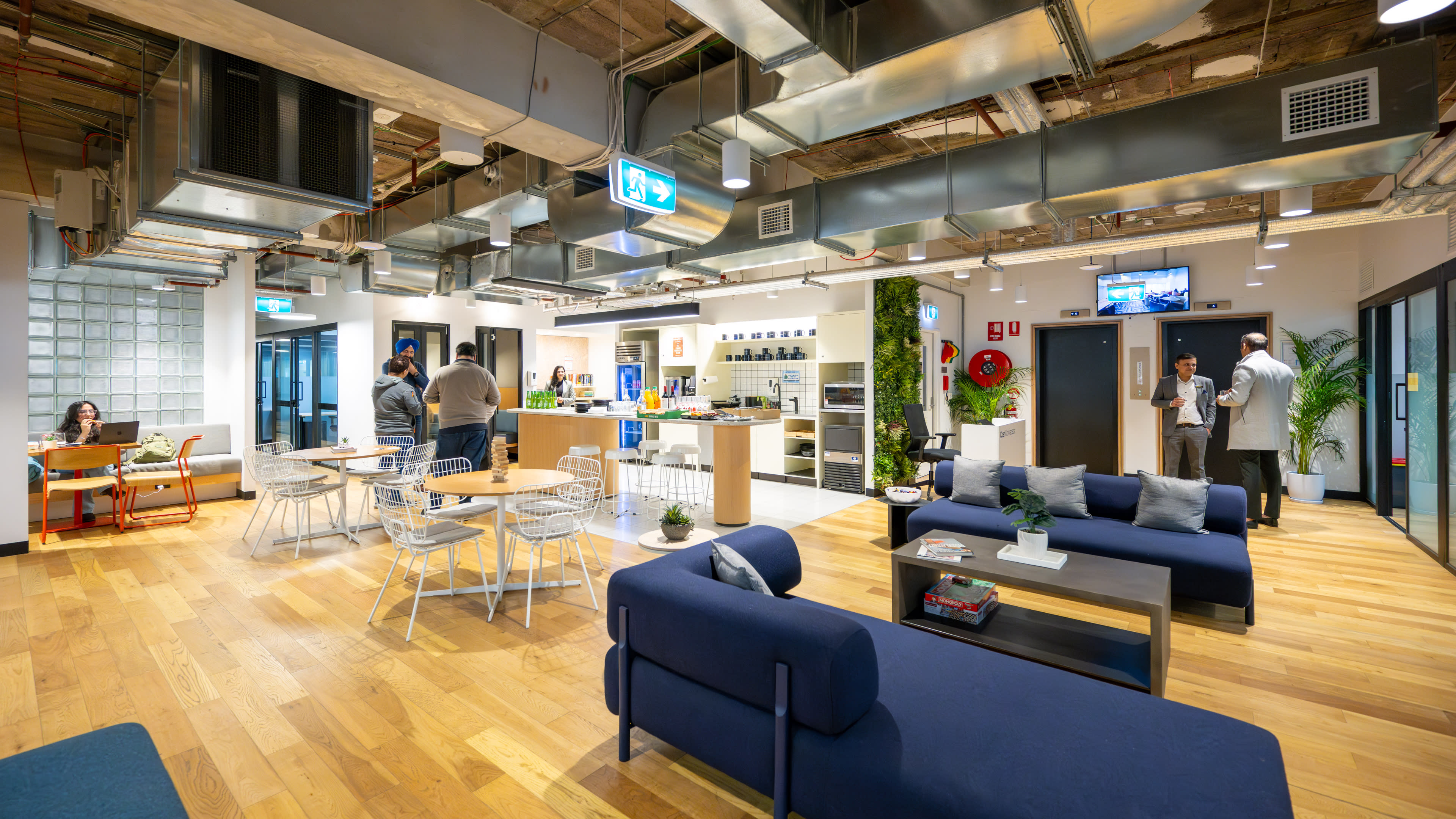 A modern office interior with a mix of seating areas, a kitchen space, and people engaged in various activities.