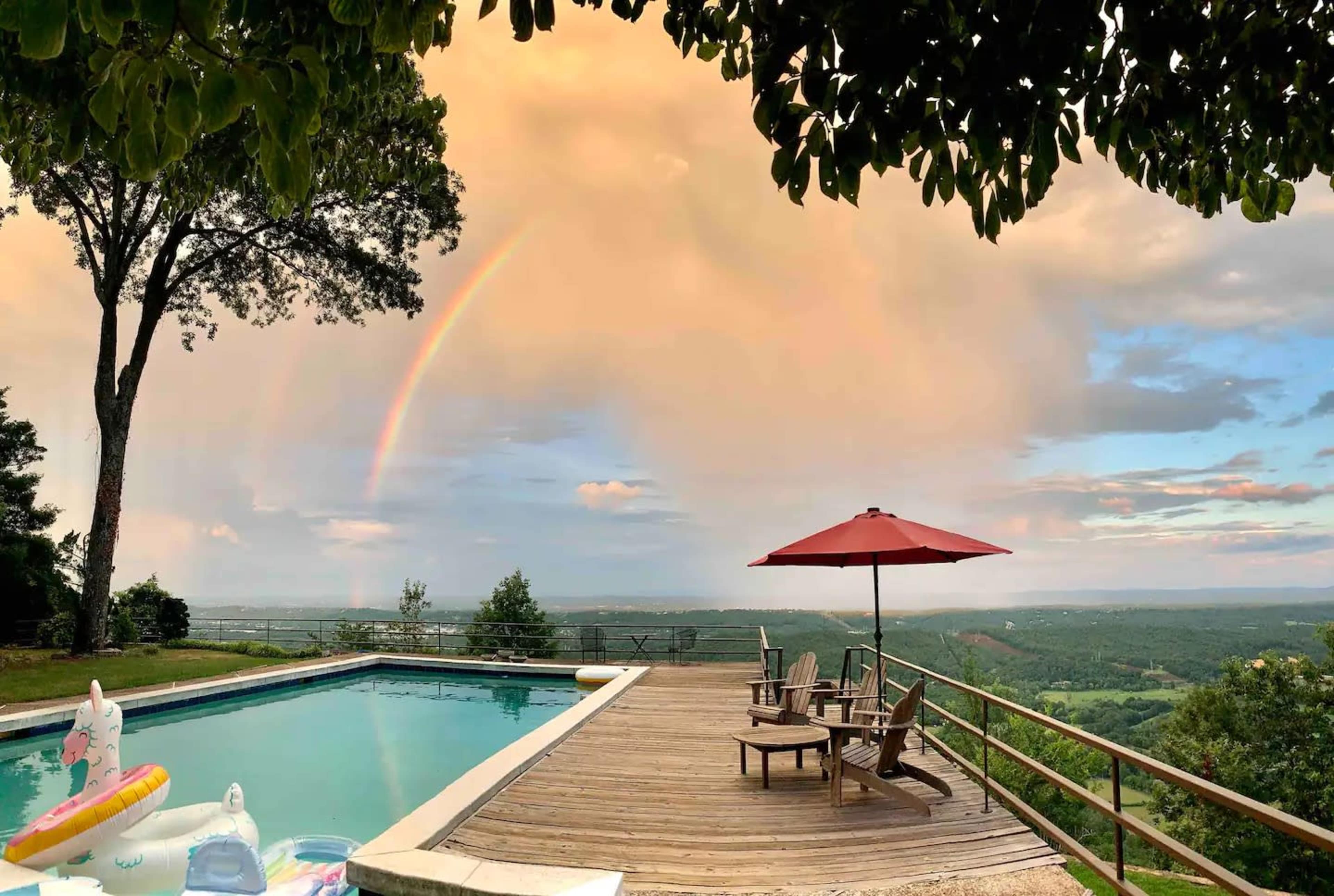 A swimming pool with wooden decking overlooks a landscape featuring a rainbow in the sky and a red patio umbrella.