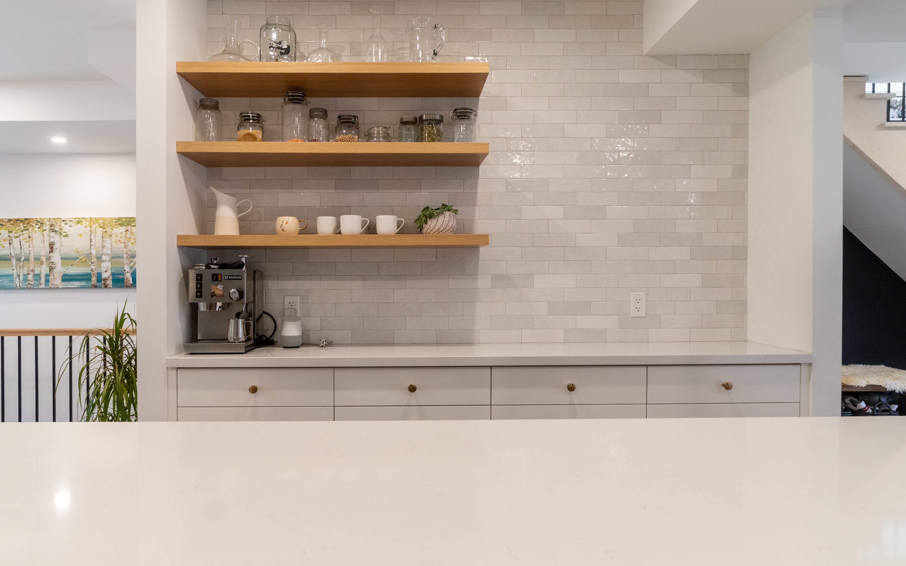 A modern kitchen features a sleek countertop with drawers below and floating wooden shelves displaying glassware and ceramic dishes on a tiled wall.