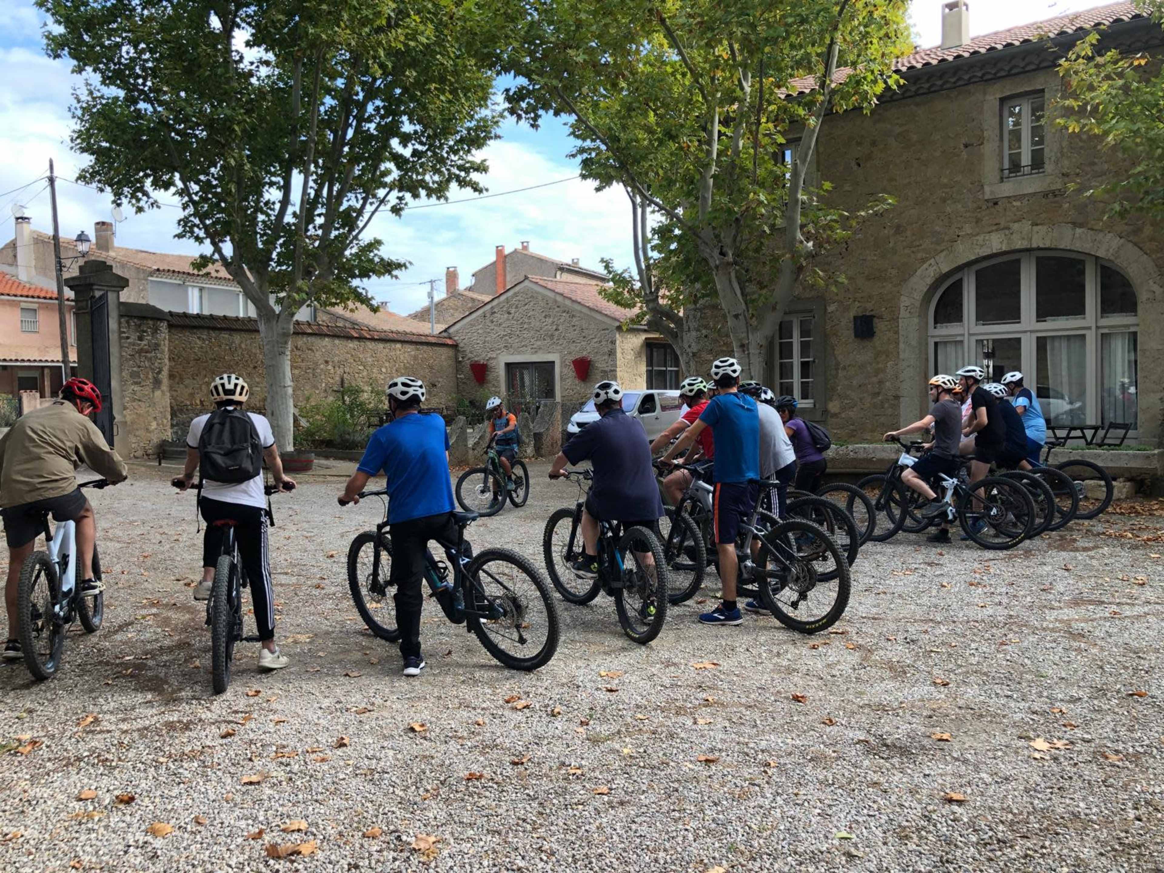A group of cyclists wearing helmets gathers around their bikes in a gravel courtyard next to a stone building.