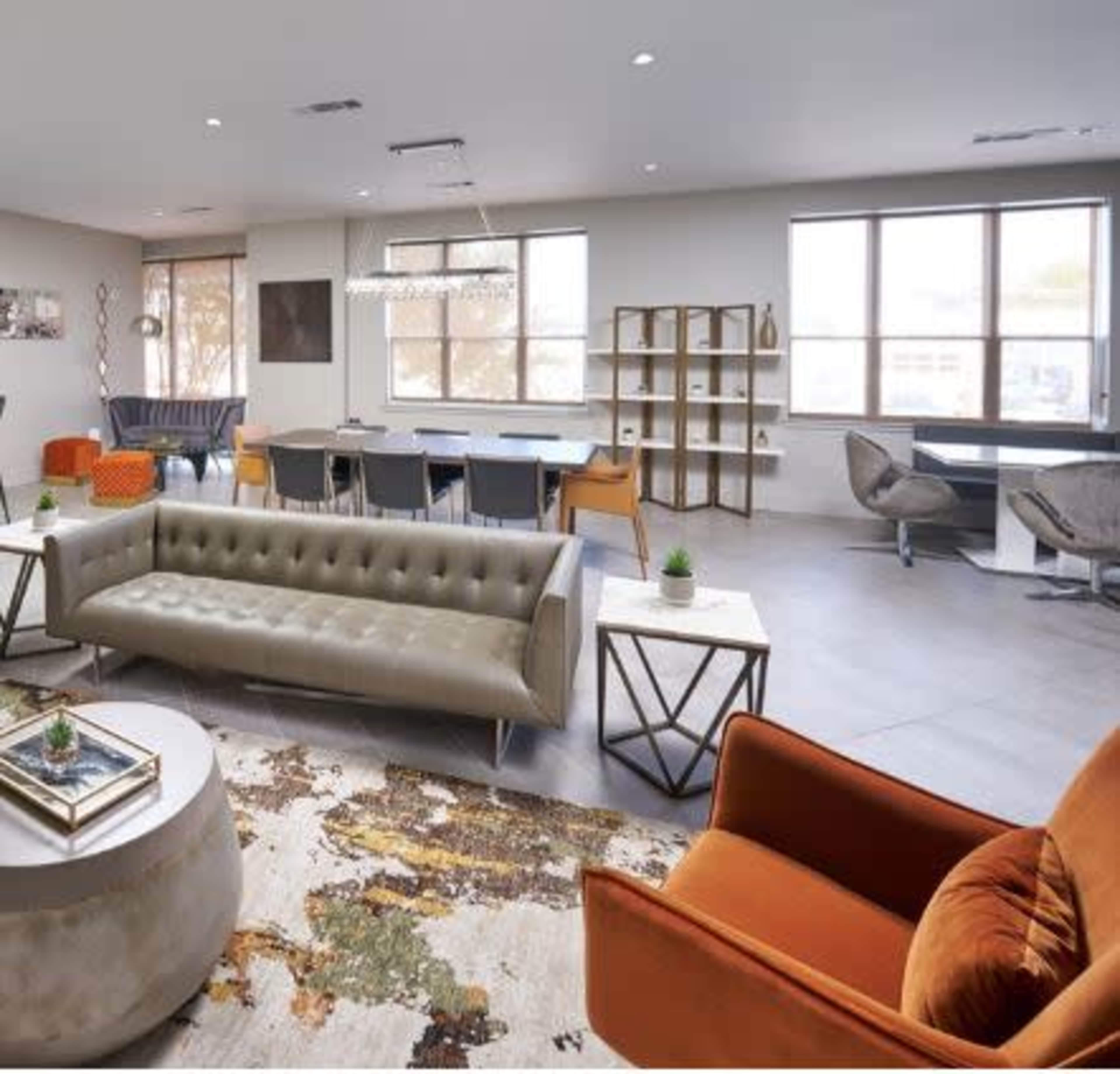 A modern lounge area with a gray sofa, orange chairs, a round coffee table, and large windows allowing natural light.