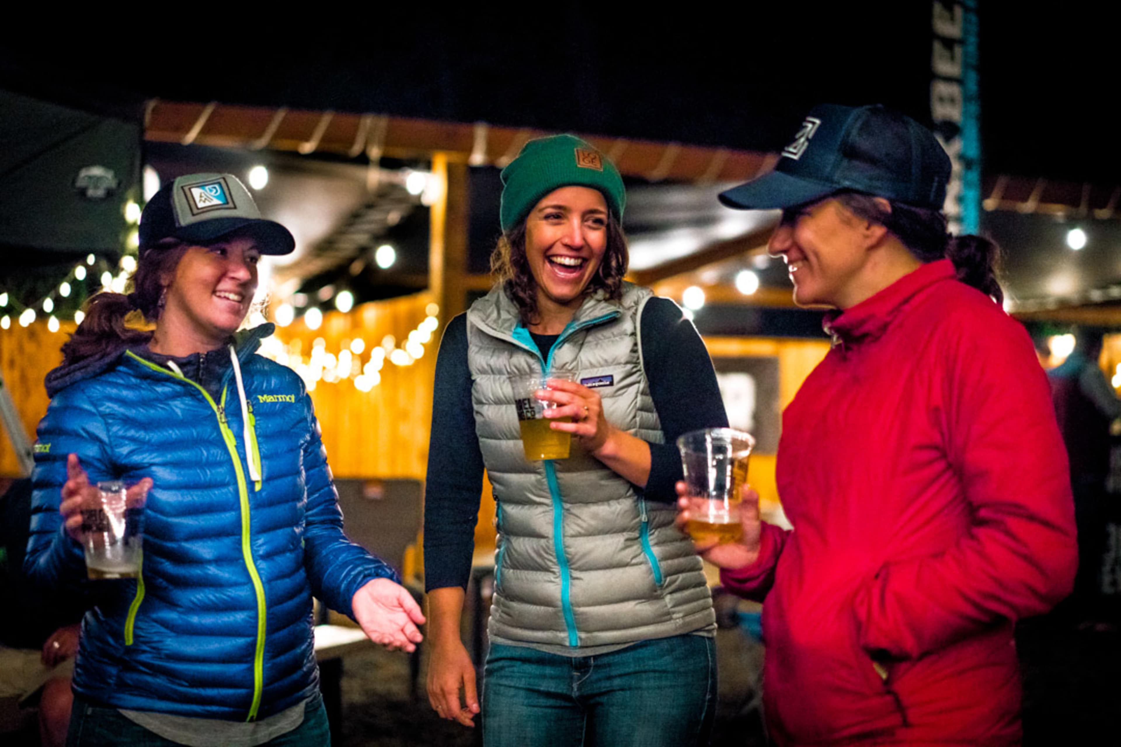 Three women are laughing and sharing drinks at an outdoor gathering under warm lights.