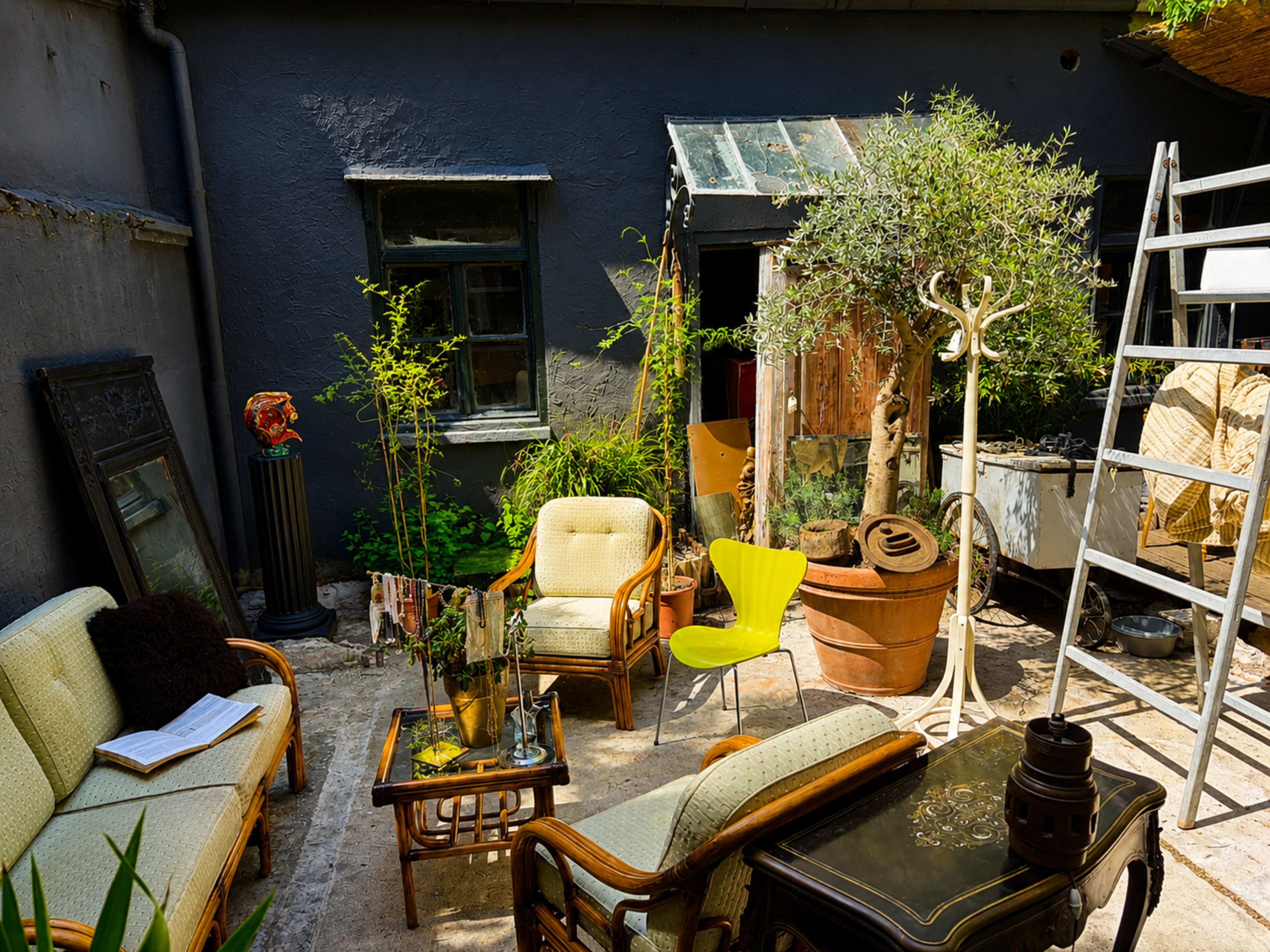 The image shows a small courtyard featuring vintage furniture, potted plants, and a ladder against a gray wall.